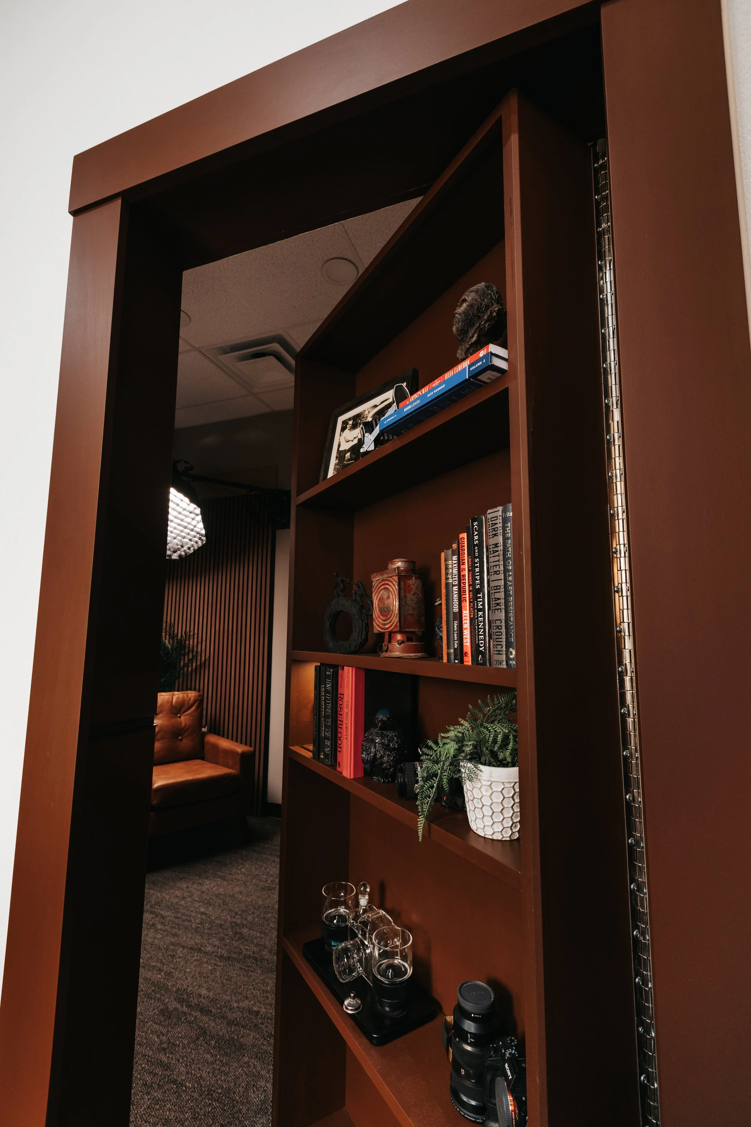 A tall wooden bookshelf with several shelves. The top shelf has a framed photo, a black furry object, and some books. Middle shelves contain more books, a potted fern, and glassware. The background reveals a cozy room with a brown leather armchair and a dark wall with vertical lines.