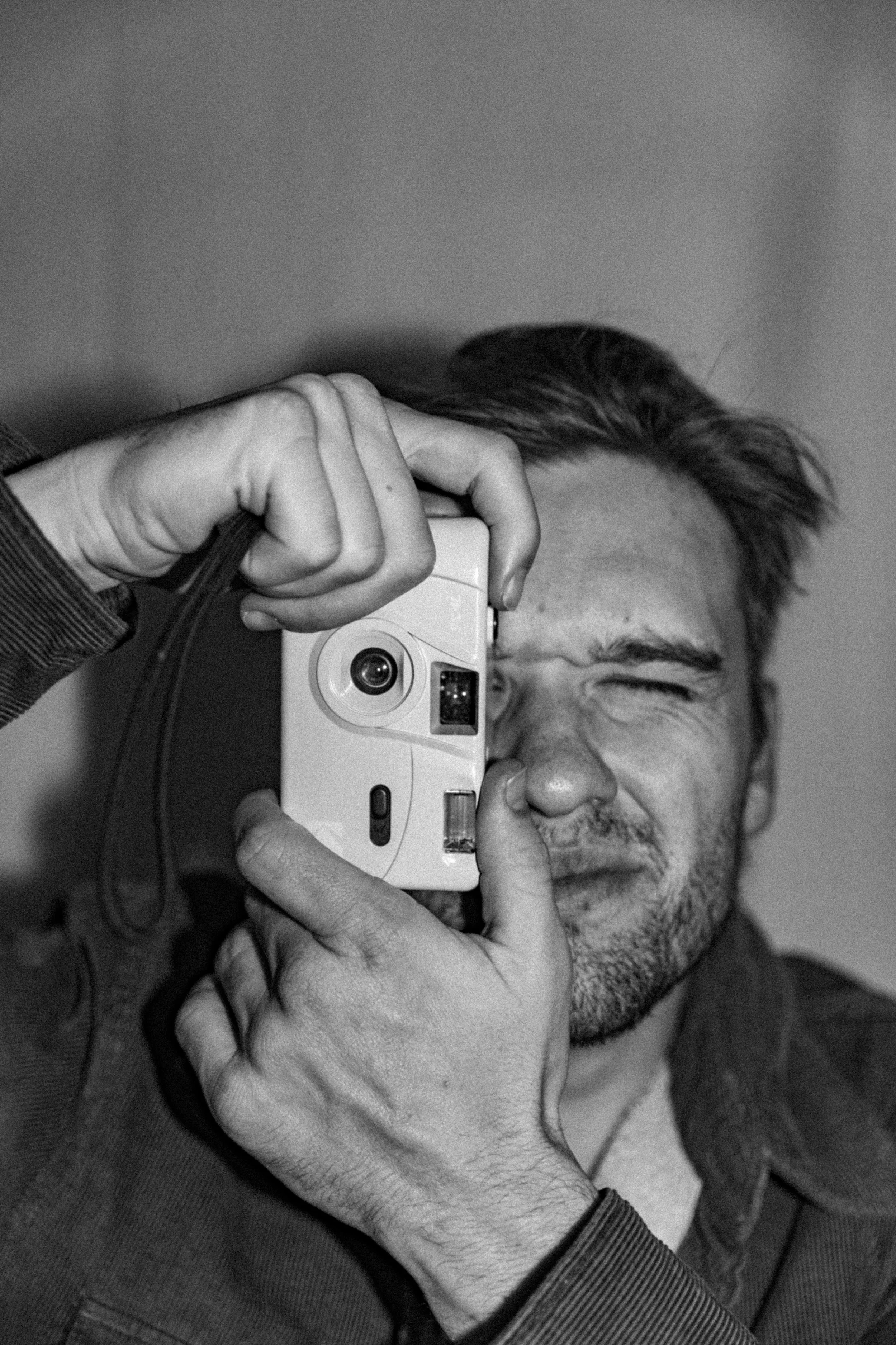 A man with short hair, a beard, and a joyful expression is taking a selfie while holding a camera. His eyes are closed, and he appears to be smiling. The background is plain and the photo is in black and white.