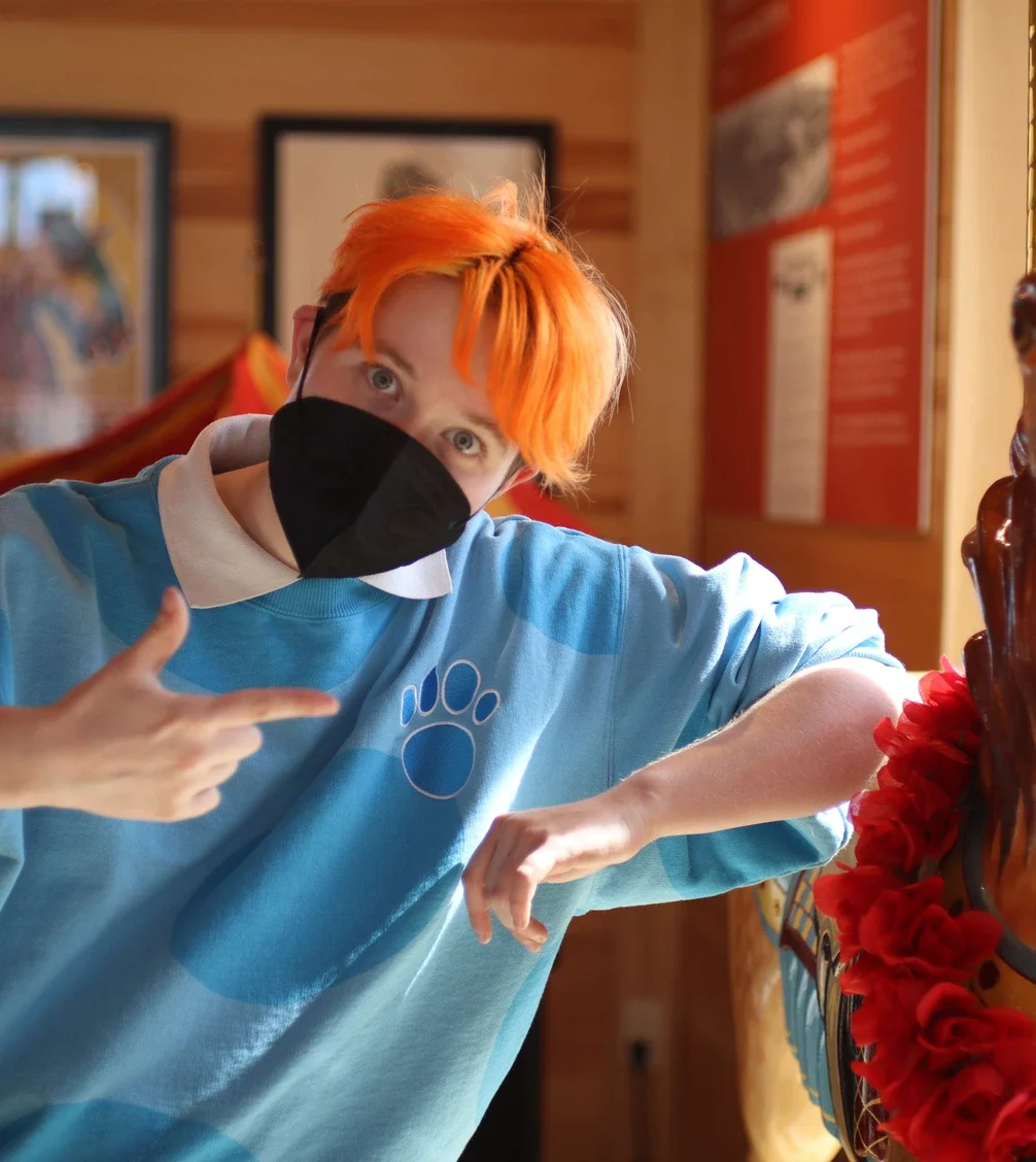 Person with bright orange hair wearing a black face mask and light blue shirt with a paw print pointing at their armpit, inside a wooden room with framed pictures and posters on the walls.