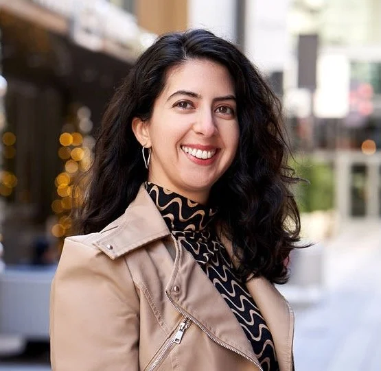 Headshot of Catarina, a light-skinned Latiné woman with dark brown wavy hair, a faux leather jacket, and a big smile in front of a blurred cityscape.