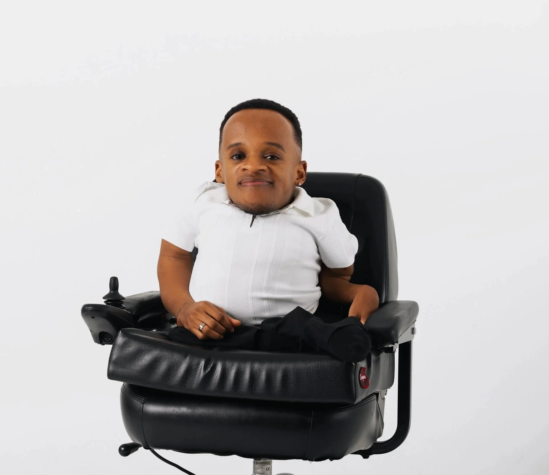 Photo of Winston, a black man, looking sharp while in his power chair looking at the camera with a smile. He is wearing a white, collared shirt and black slacks in front of a cream colored backdrop.