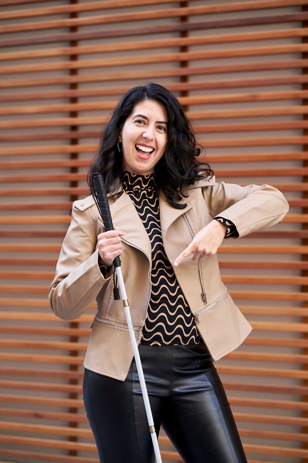 Photo of Catarina, a light skinned Latine woman, wearing a tan/khaki trench coat, holding her blindness cane and pointing to it. Behind her is a wood paneled wall that almost looks like it has shutters because of the wood style.