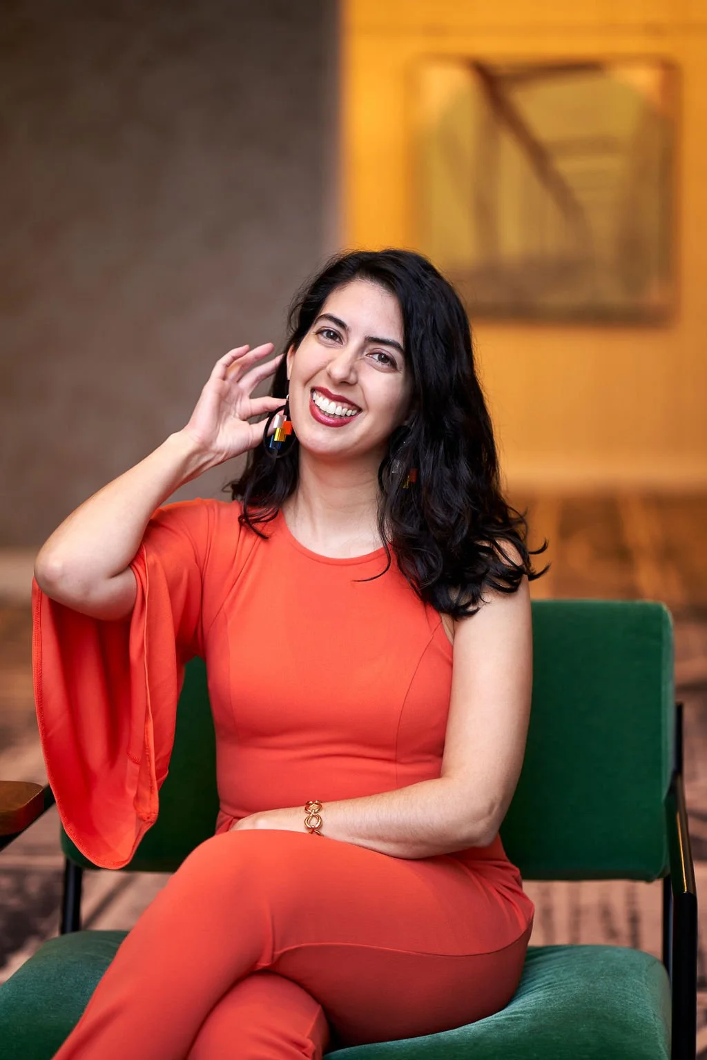 Catarina, a light-skinned Latine woman with dark brown wavy hair, is  wearing a bright orange jumpsuit and smiling at the camera. Her right hand is raised, lightly touching a gold and red earring while her left arm rests on her lap. Her legs are cros
