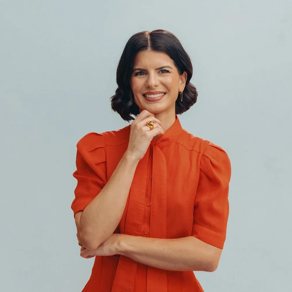 Photo of Margaux, a white woman with her hair in a beautiful bob. She is looking at the camera smiling and wearing a vibrant orange collared shirt with slightly puffed sleeves. Her right hand is bent up under her chin and she's holding a pencil.