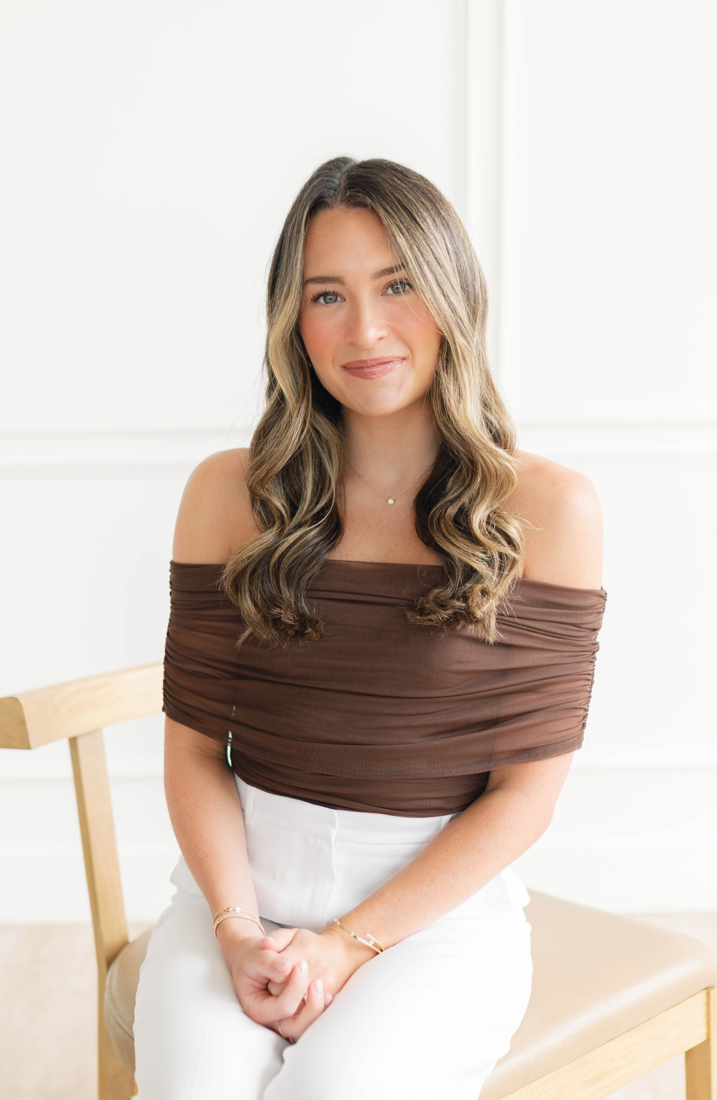A woman with long, wavy brown hair and light skin, sitting on a light wooden chair, wearing an off-the-shoulder brown top and white pants, smiling softly.
