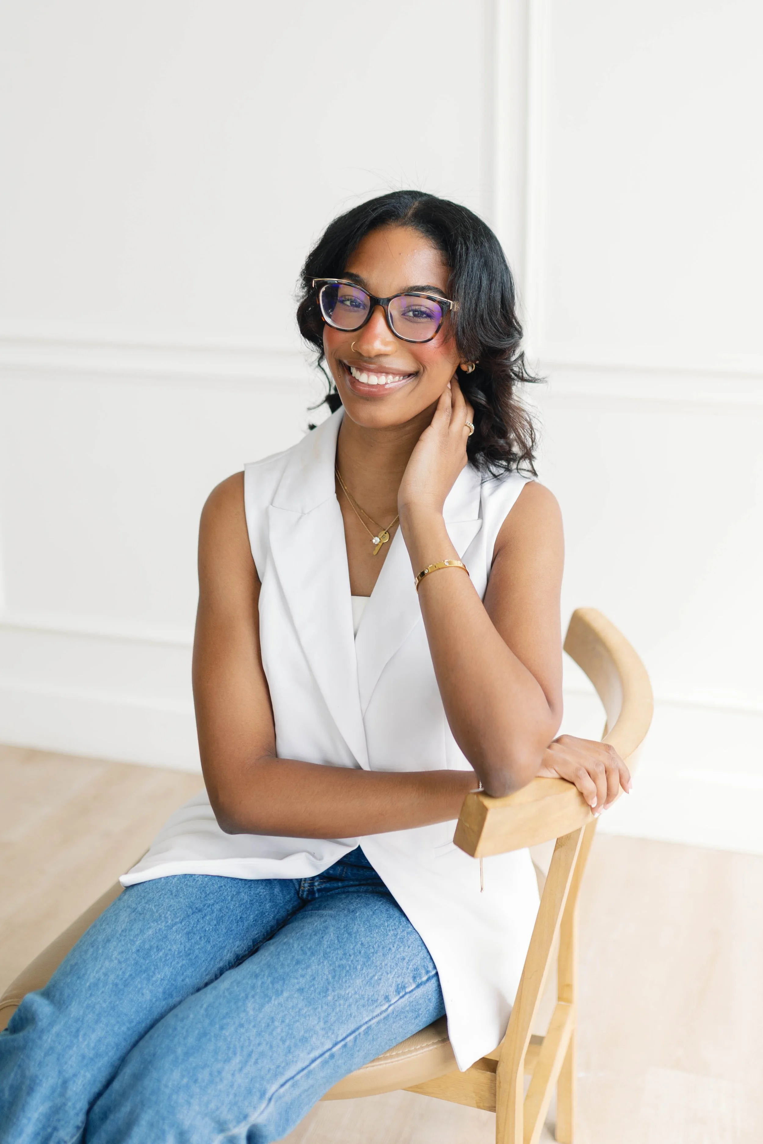 A woman smiling while sitting on a wooden chair, wearing a white sleeveless blazer, blue jeans, glasses, and jewelry, with a plain white wall in the background.
