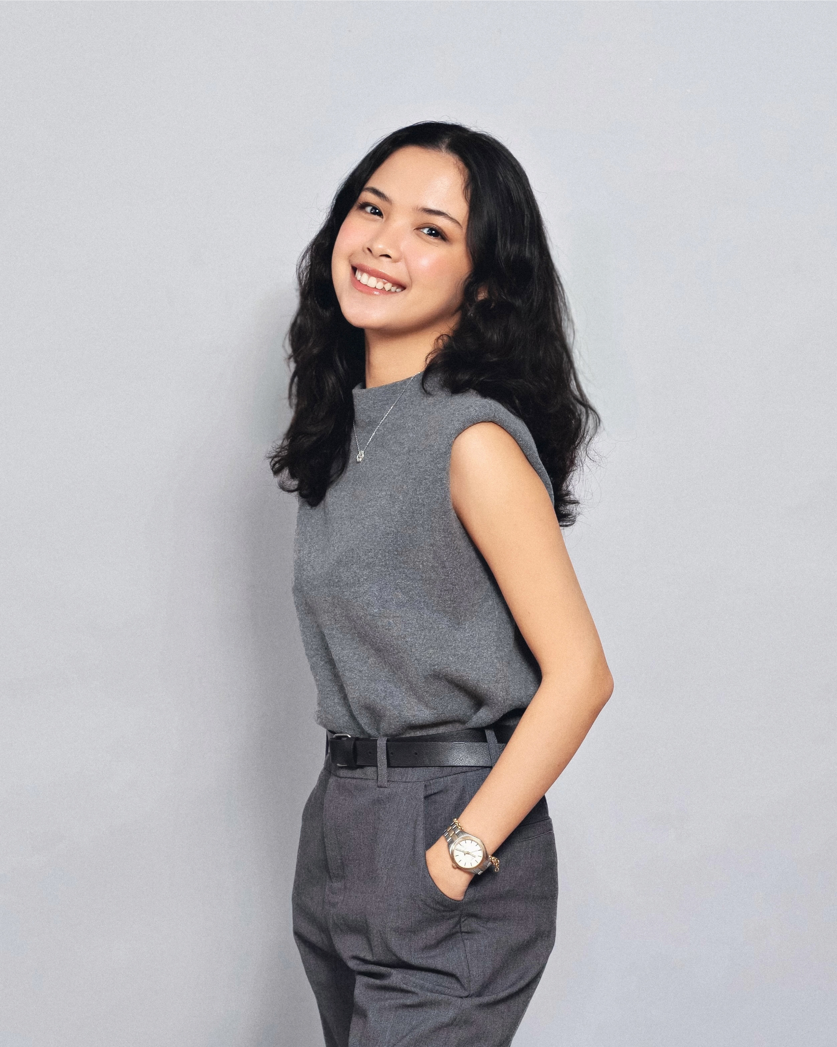 A young woman with dark, wavy hair smiling and posing with her hand in her pocket, wearing a sleeveless gray top, gray pants, a wristwatch, and a necklace, standing against a plain light gray background.