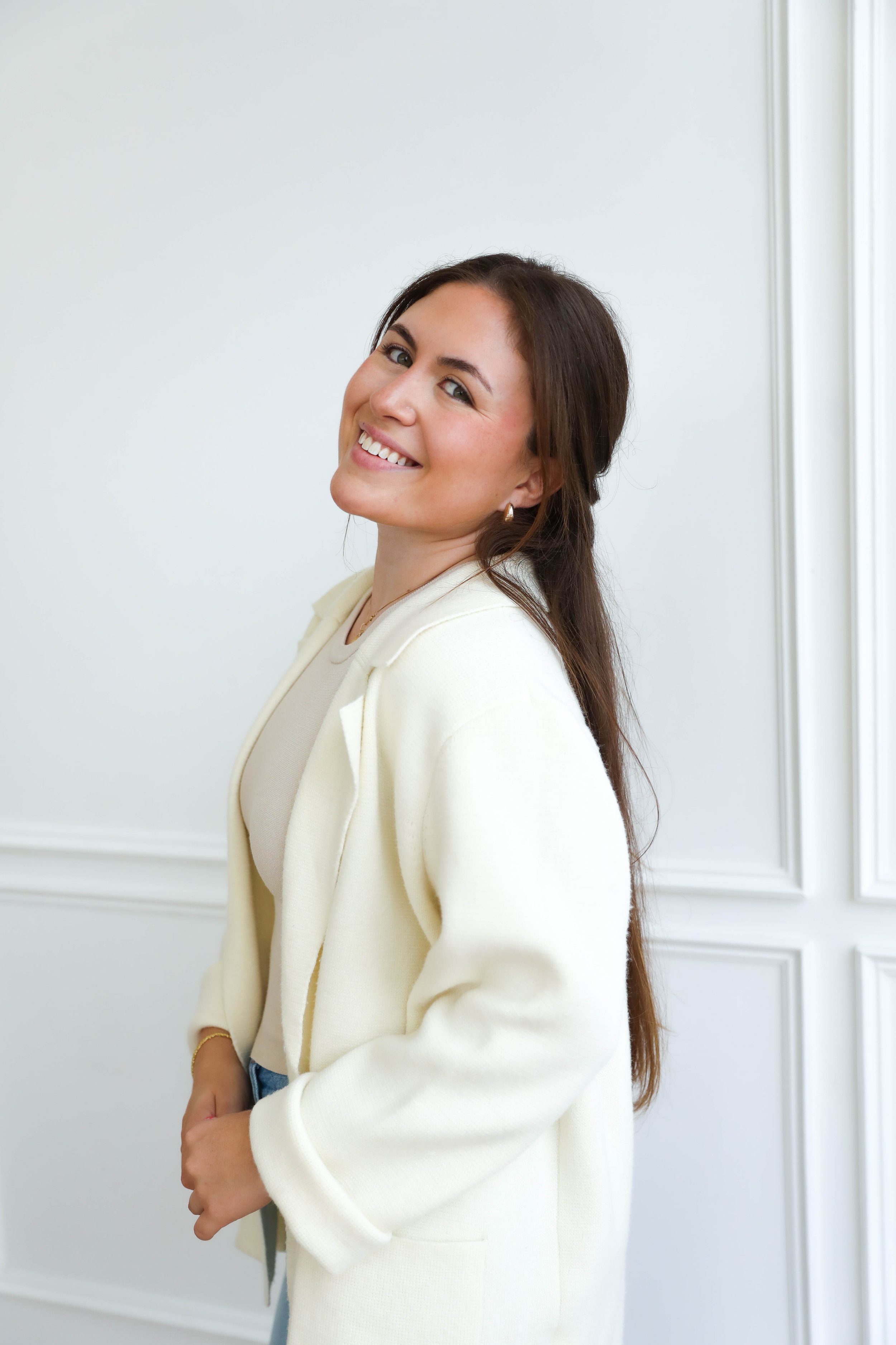 A young woman with long brown hair smiling and looking at the camera, standing against a white wall with decorative paneling, wearing a cream-colored jacket and earrings.