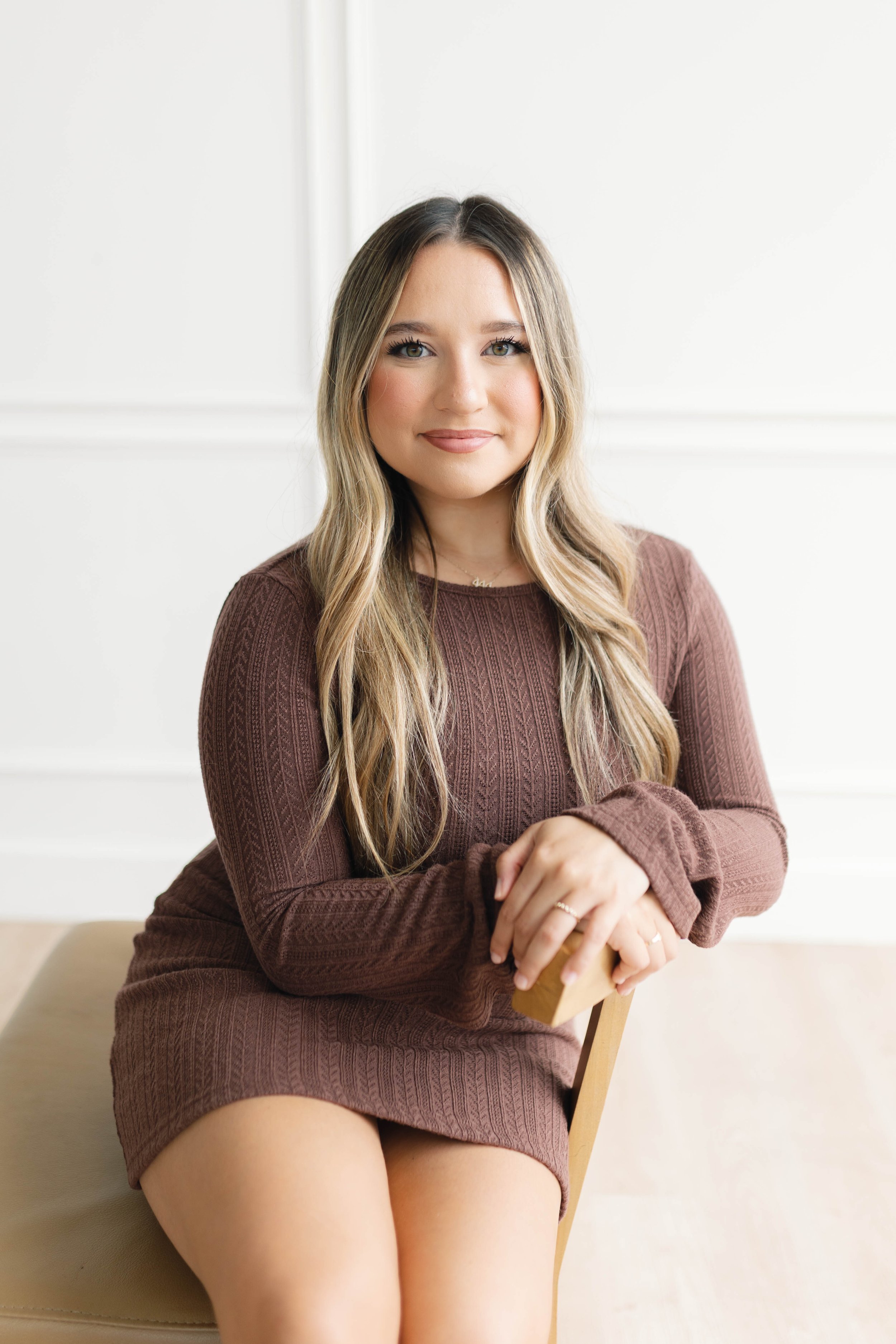 Young woman with long blonde hair sitting on a beige chair, wearing a brown long-sleeve dress, smiling, in a bright room with white walls.