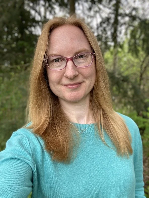 A woman named Angela has on a blue sweater and is gently smiling. She has white skin, pink glasses, green eyes and natural red hair.