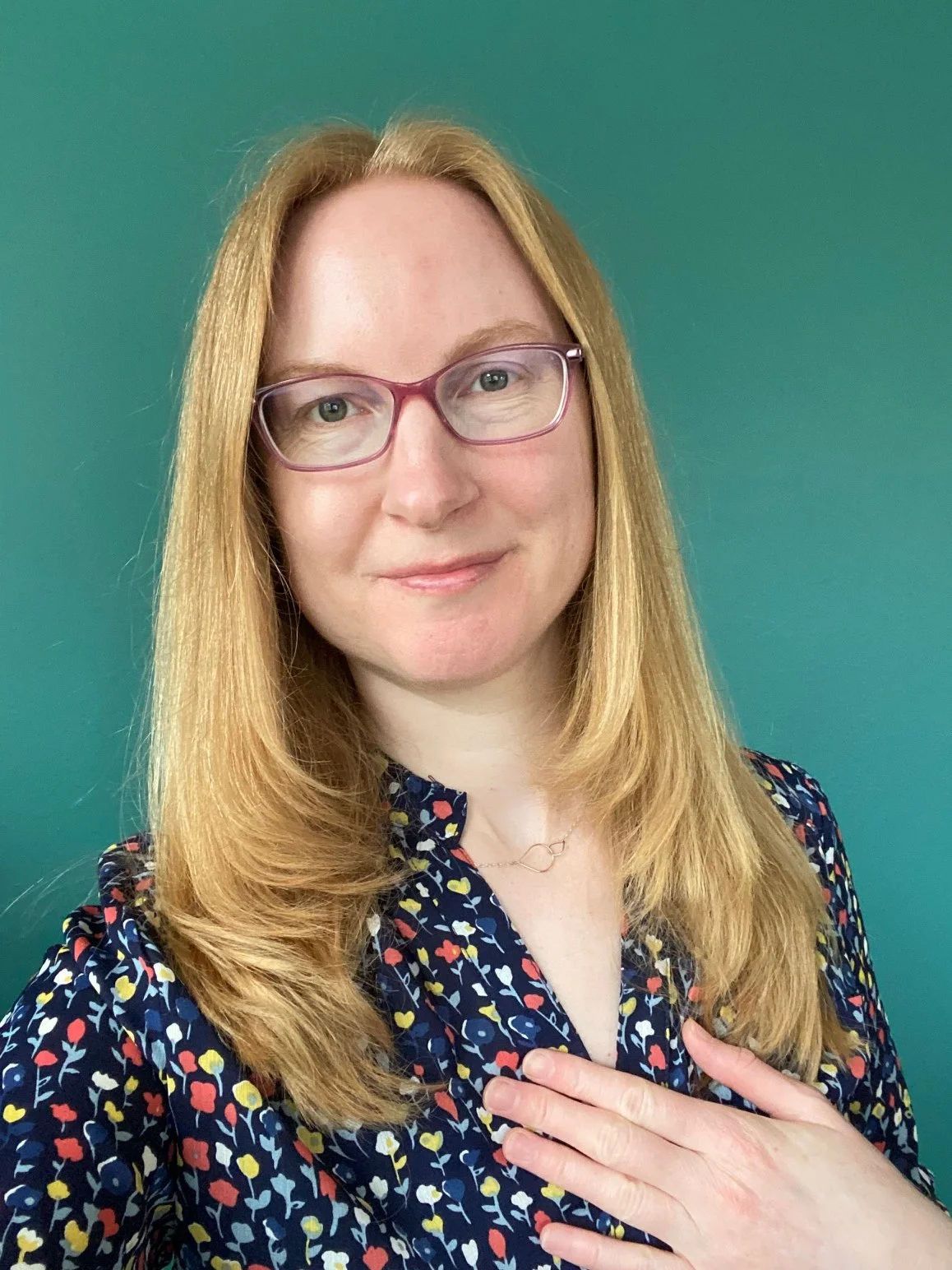 A woman named Angela is gently smiling, while wearing glasses and a floral blouse. Representing empathy and comfort for grieving pet parents.