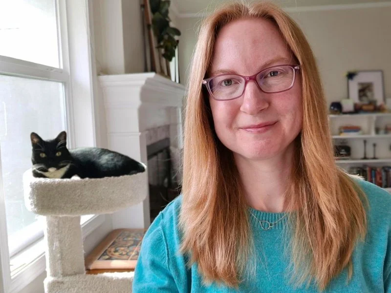 A woman is wearing a blue sweater, she is smiling with her cat behind her in a cat tree.