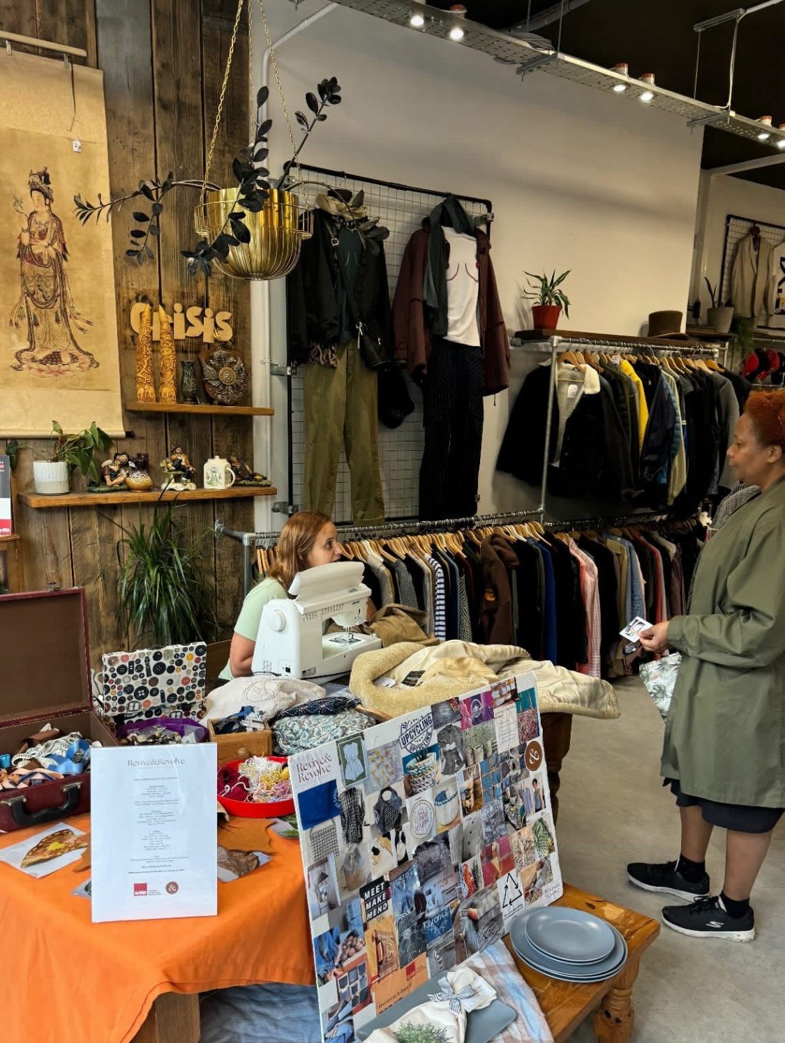 Inside a clothing store with various clothes displayed on racks and walls, a counter with a sewing machine, and a person interacting with a customer. The space is decorated with plants, and a tablecloth displays craft materials and a collage board.