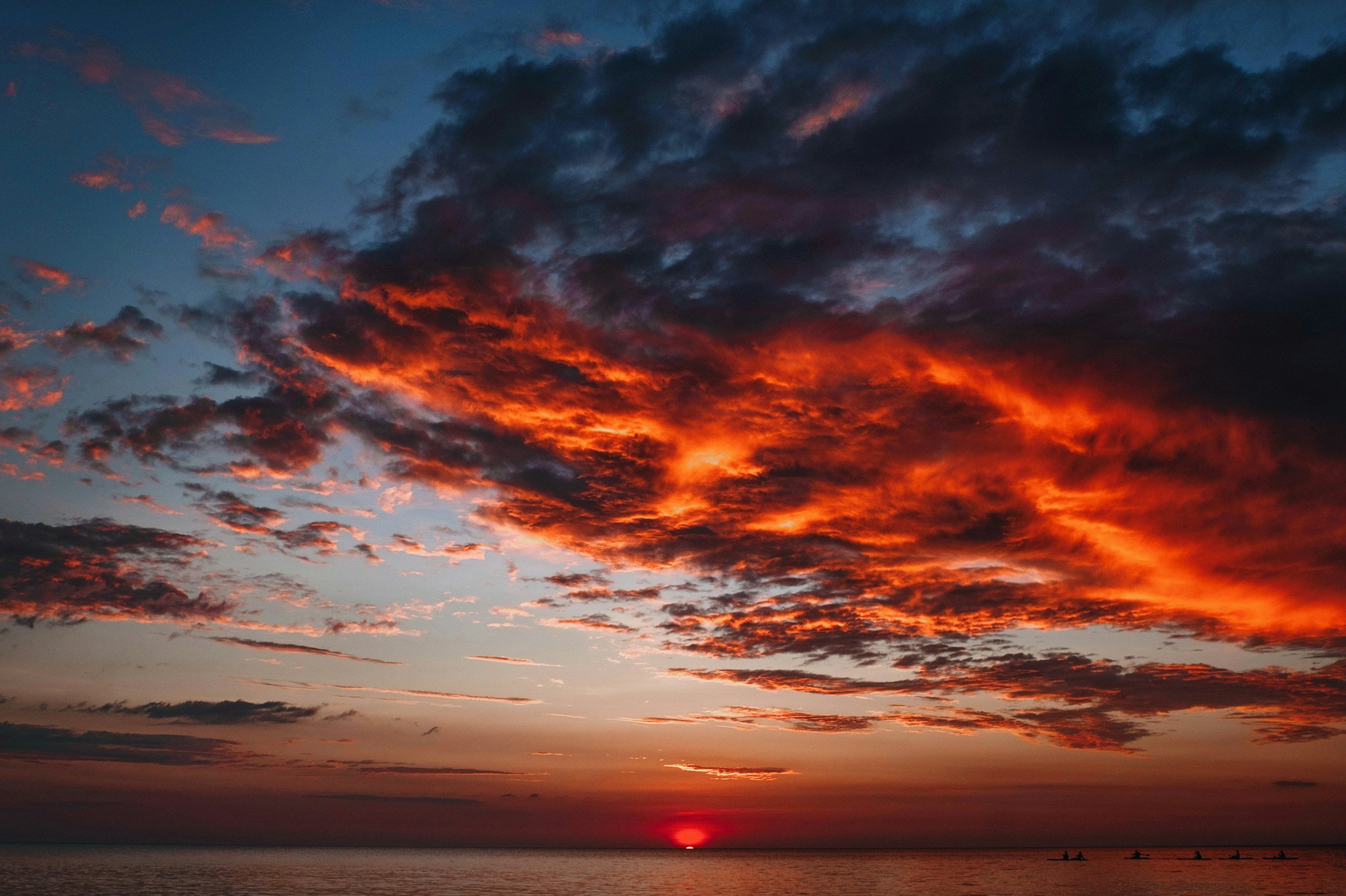 Sunset over the ocean with dark clouds illuminated by orange and red hues, small boats on the water in the distance.