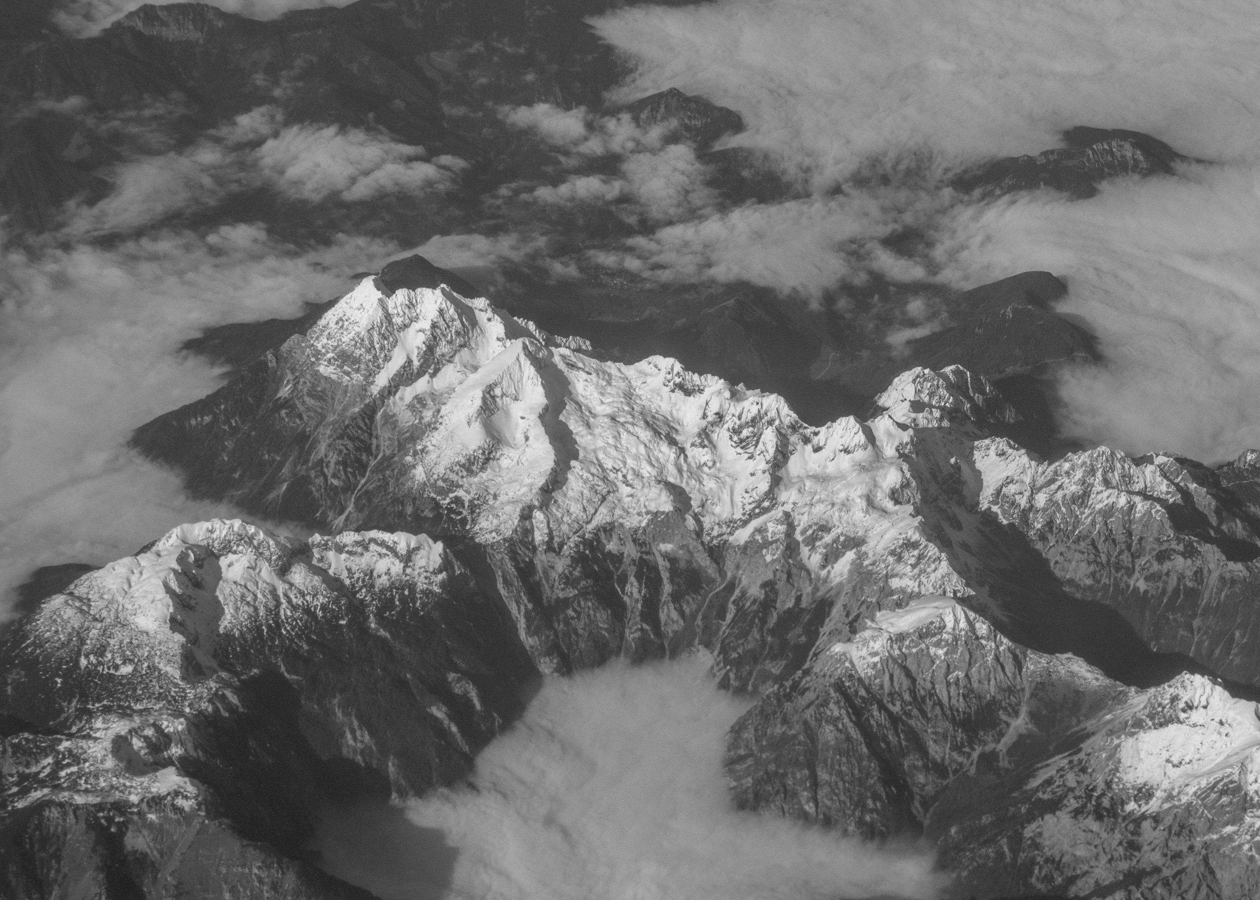 Aerial black and white photograph of snow-capped mountain range with rugged peaks, some shrouded in clouds.