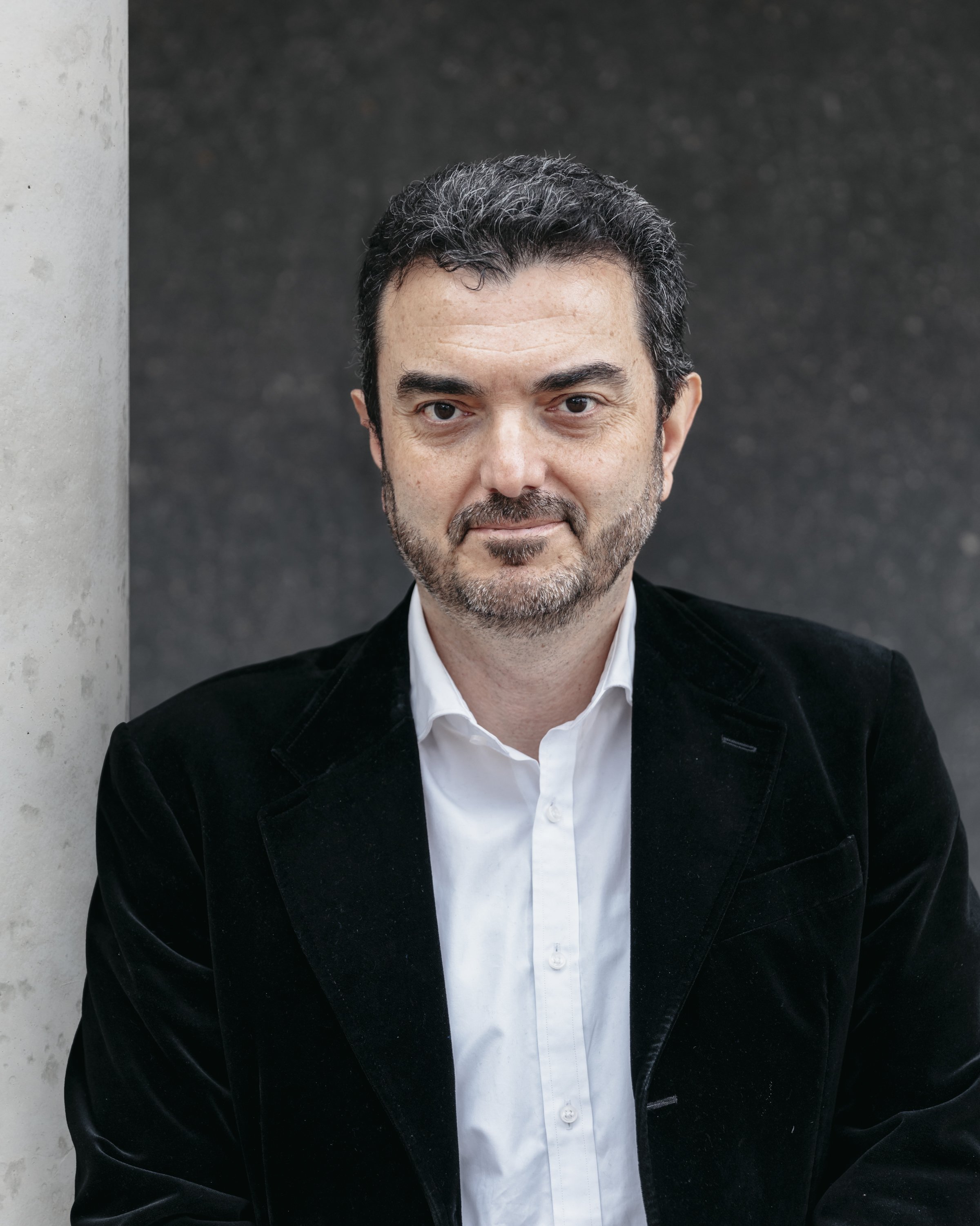 A man in a black jacket and white shirt looks warmly at the camera