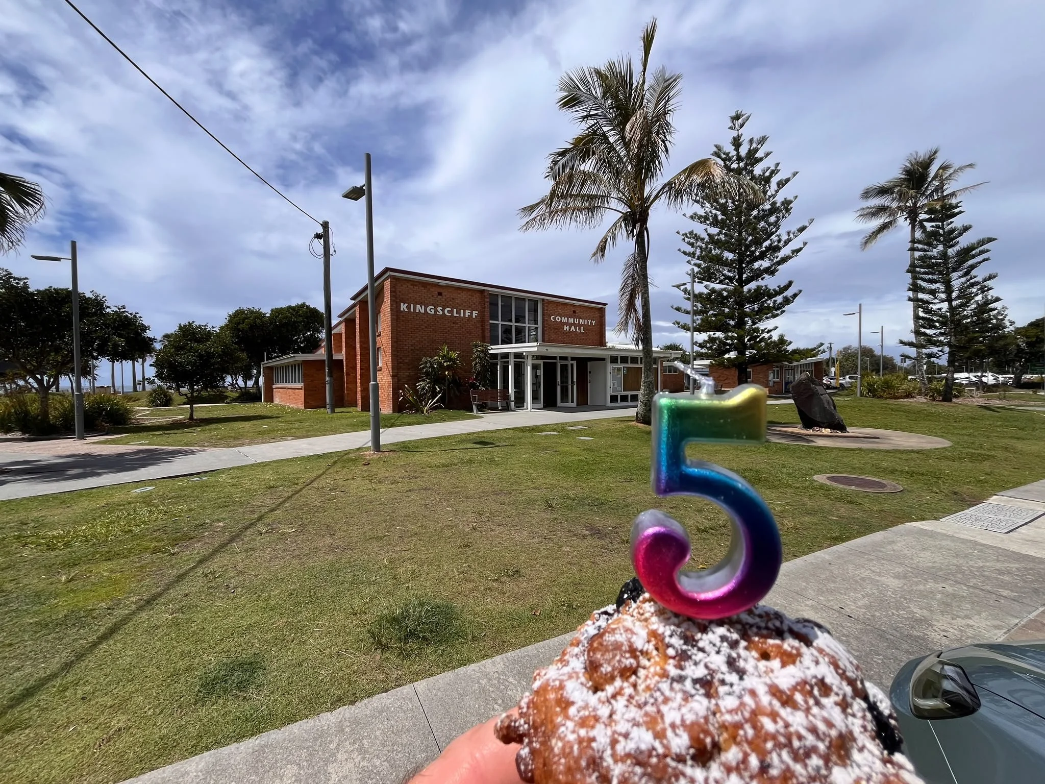A muffin with a 5 candle is held in front of a red brick community hall building surrounded by trees