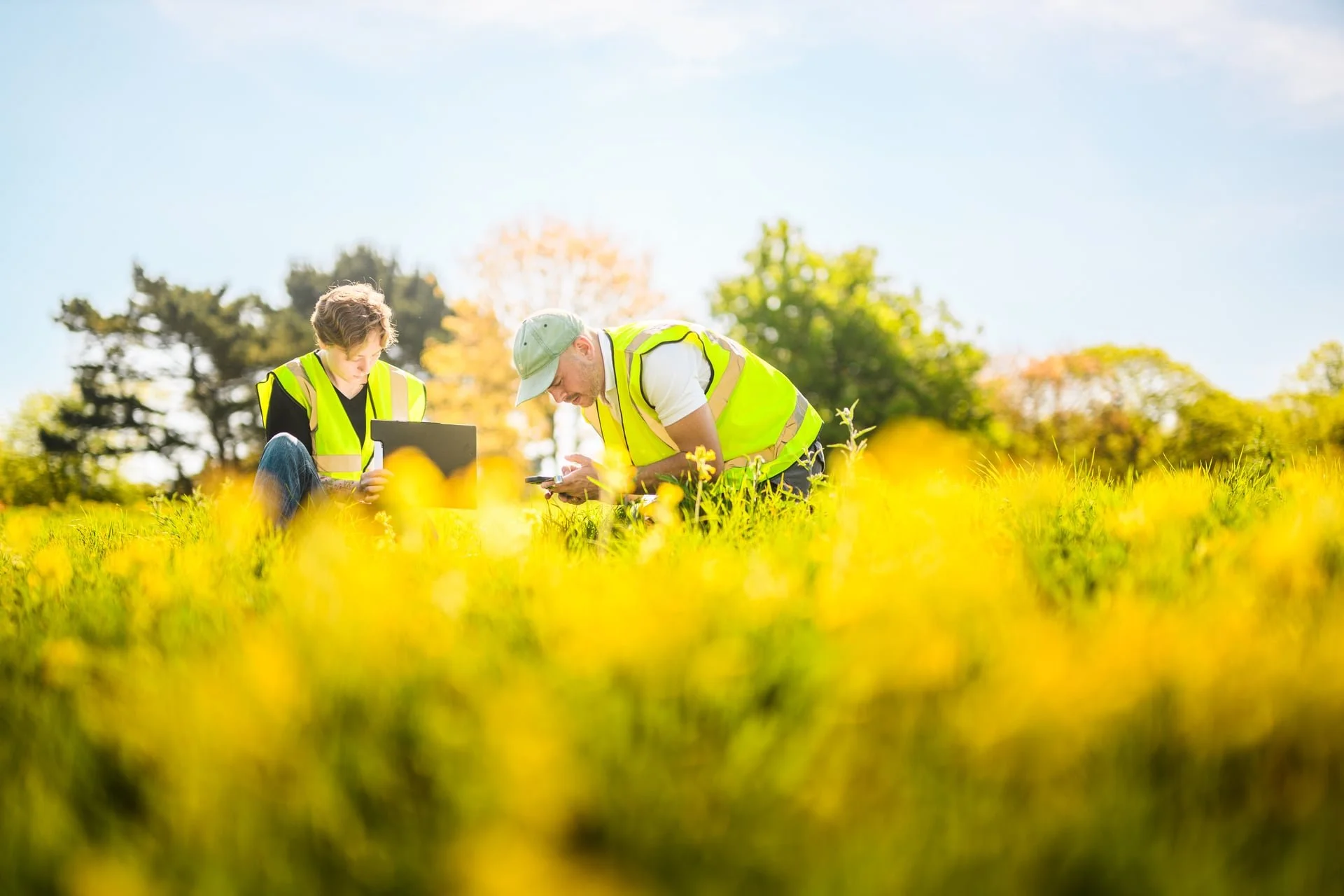 Two men in yellow safety vests working outdoors in a grassy field, examining documents or a device, with trees and a partly cloudy sky in the background.