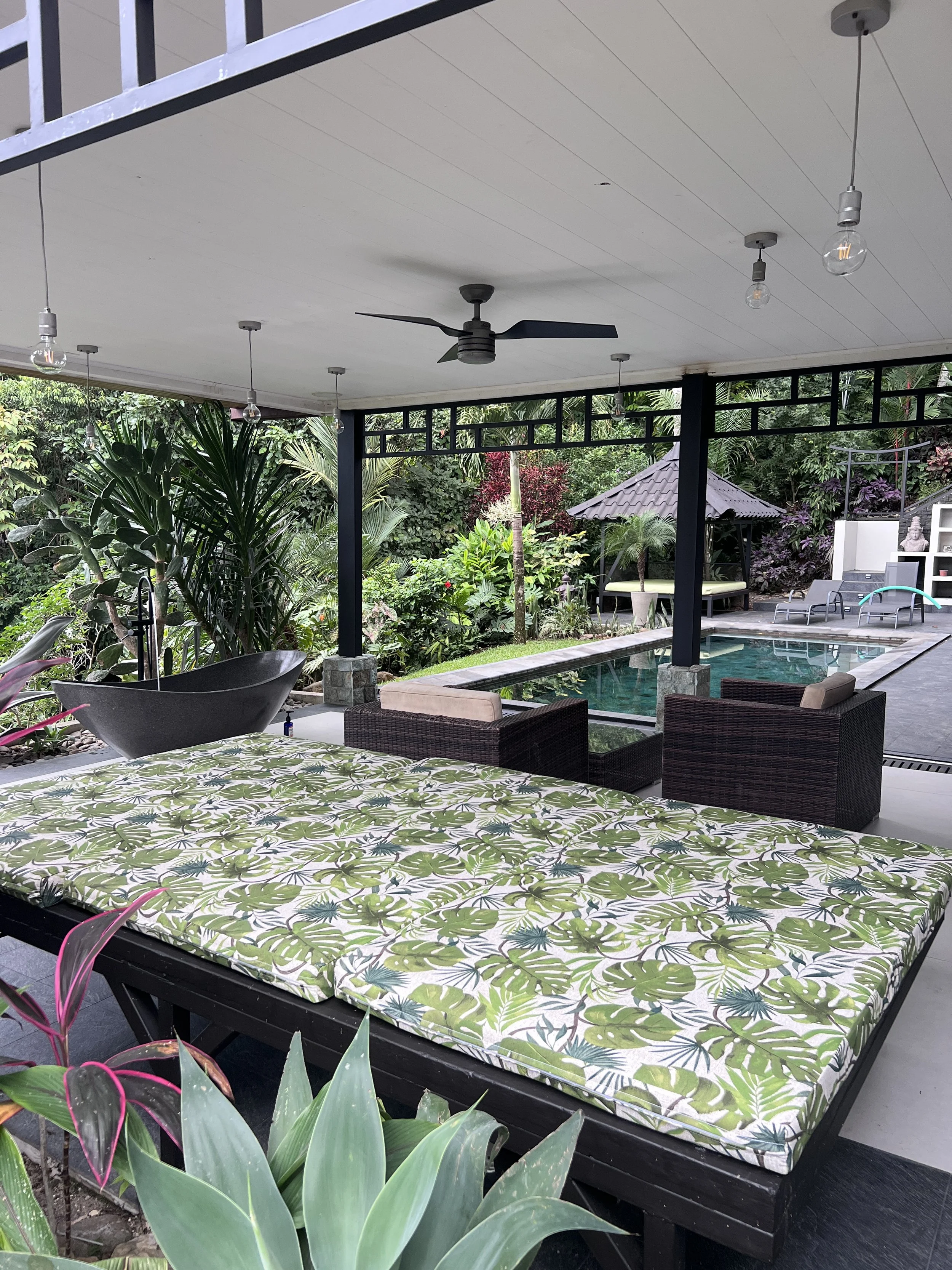 Outdoor patio area with tropical plants, a swimming pool, a shaded seating area with wicker chairs, a gazebo, hanging light bulbs, and a ceiling fan.
