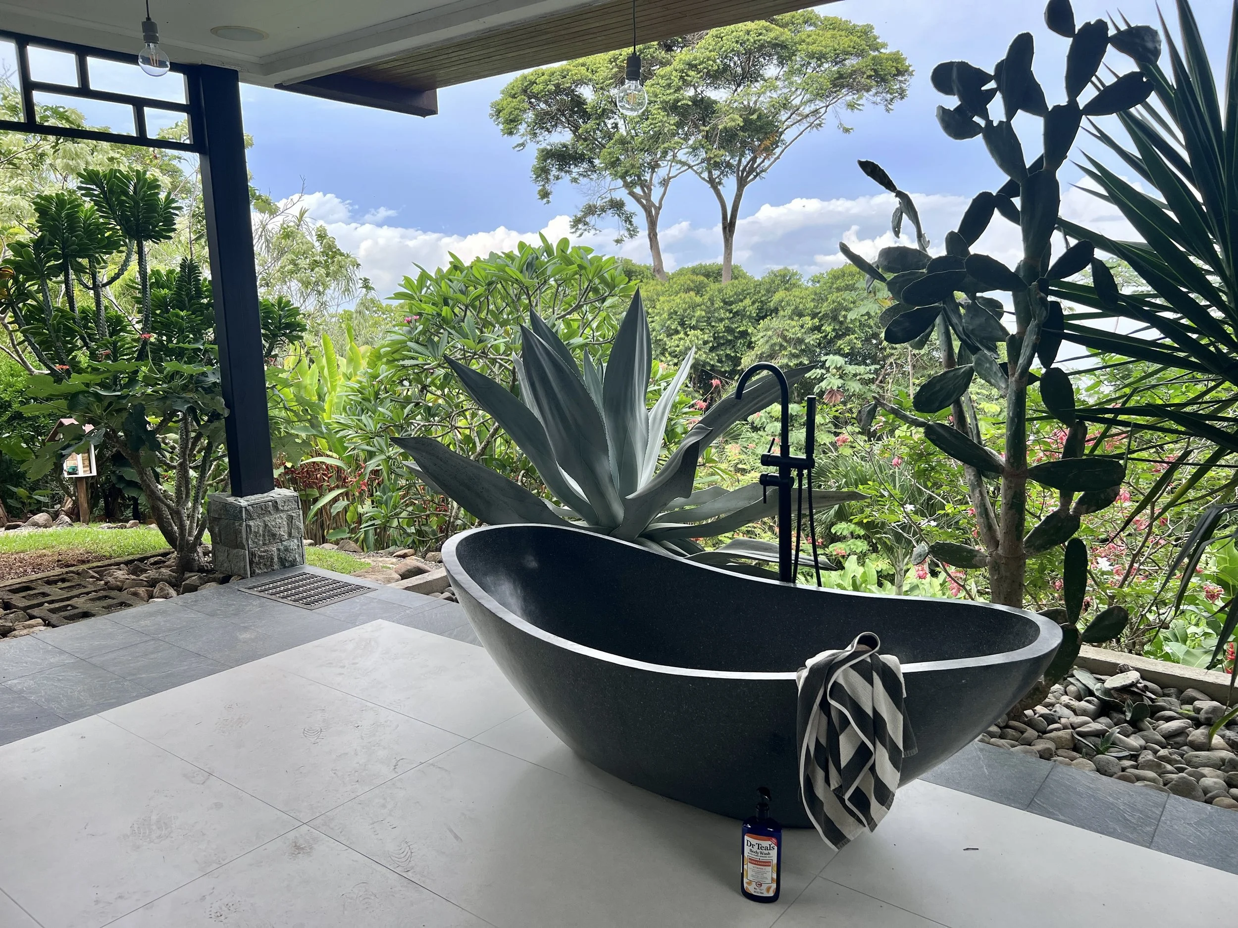 A modern outdoor shower area with a black oval-shaped bathtub and a striped towel hanging over the side, set against lush green tropical plants and trees, with large gray tiles on the floor and a view of the blue sky and trees beyond.