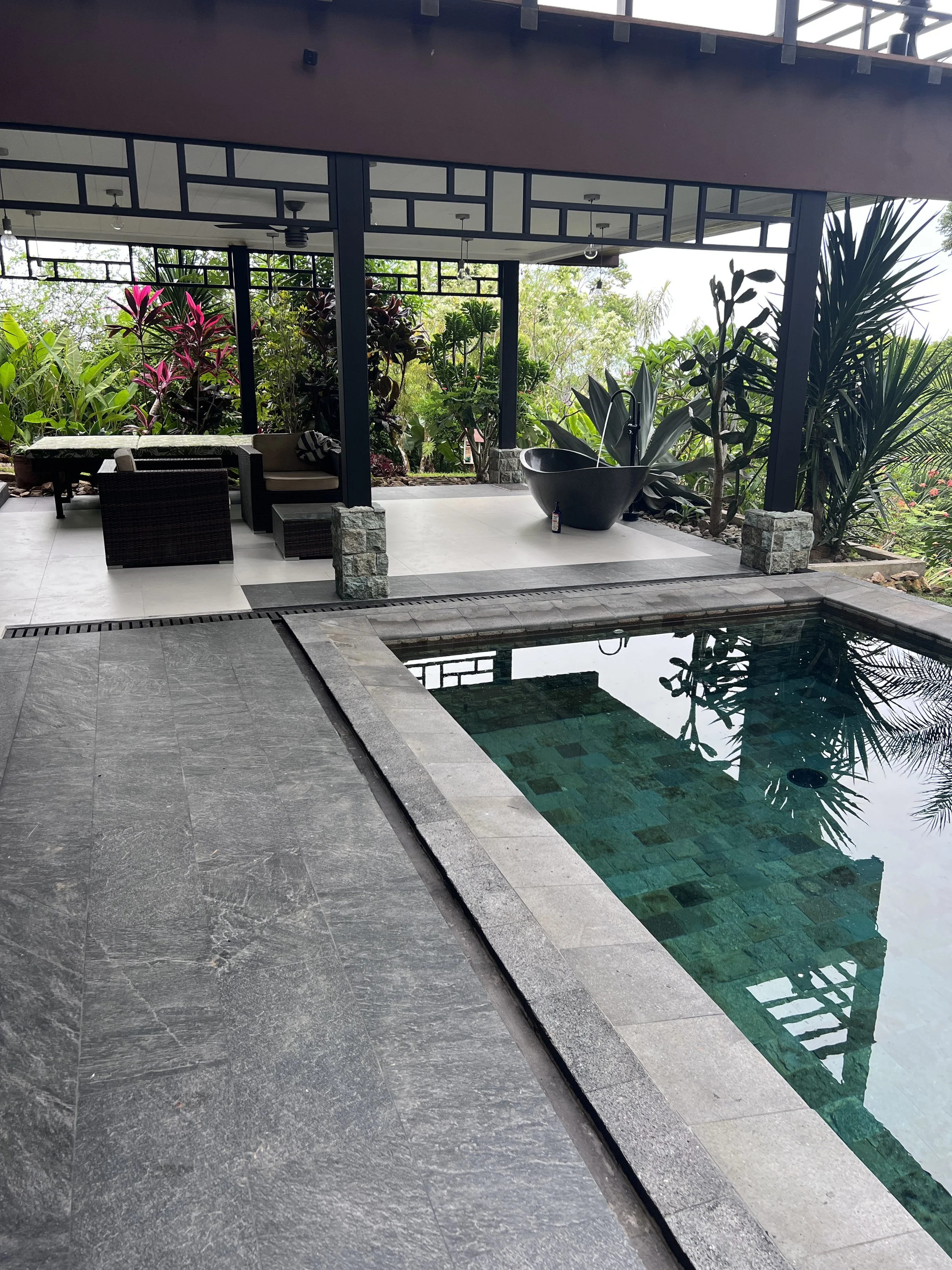 A backyard with a small rectangular swimming pool, surrounded by gray stone tiles. There is an outdoor seating area with wicker furniture and cushions, and a small black basin with water. The background has lush green plants and trees, and an upper b
