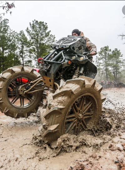 A person riding a modified all-terrain vehicle with large tires through muddy terrain with trees in the background.