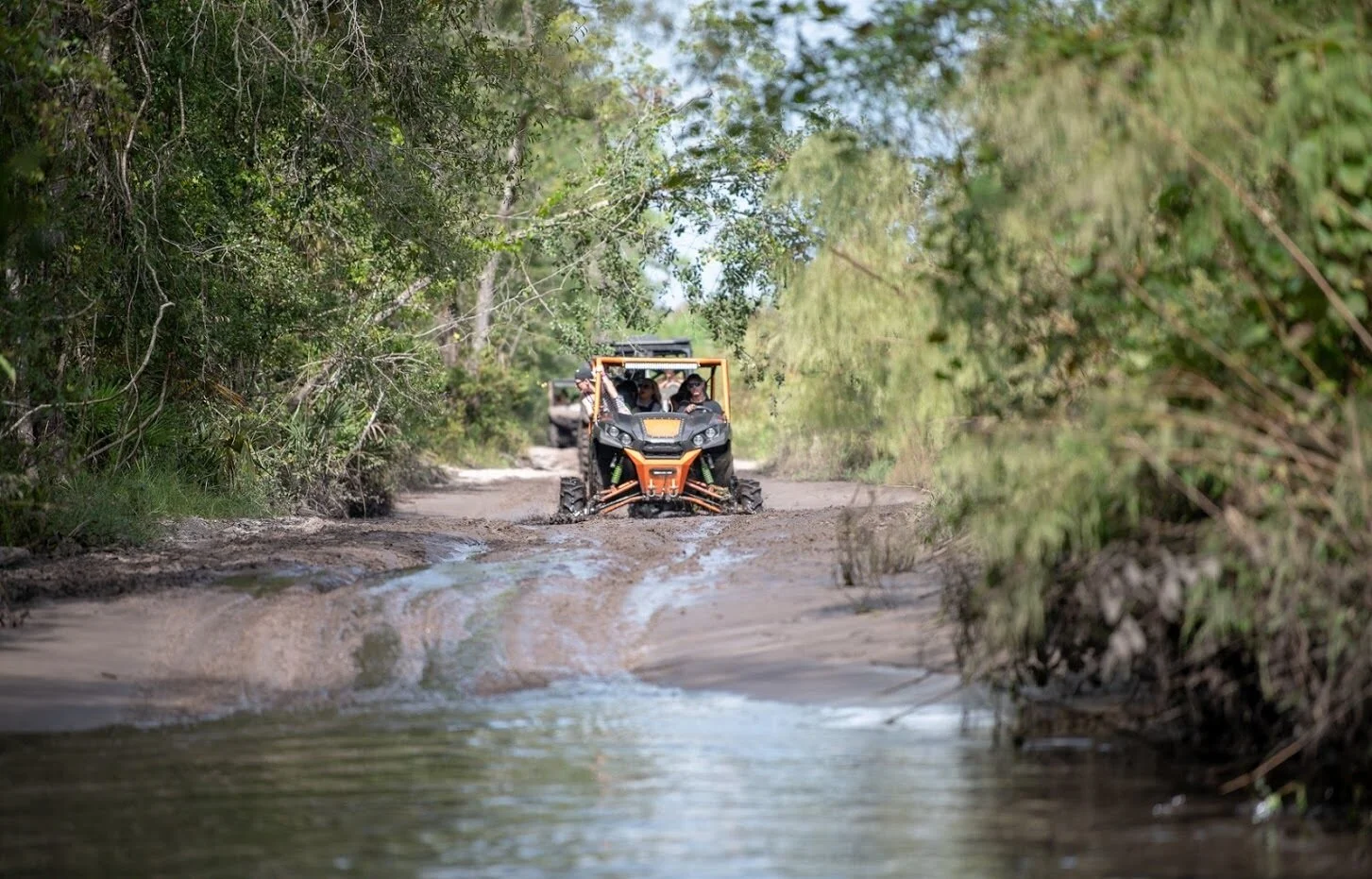 An off-road vehicle driving through a muddy trail surrounded by trees and greenery.
