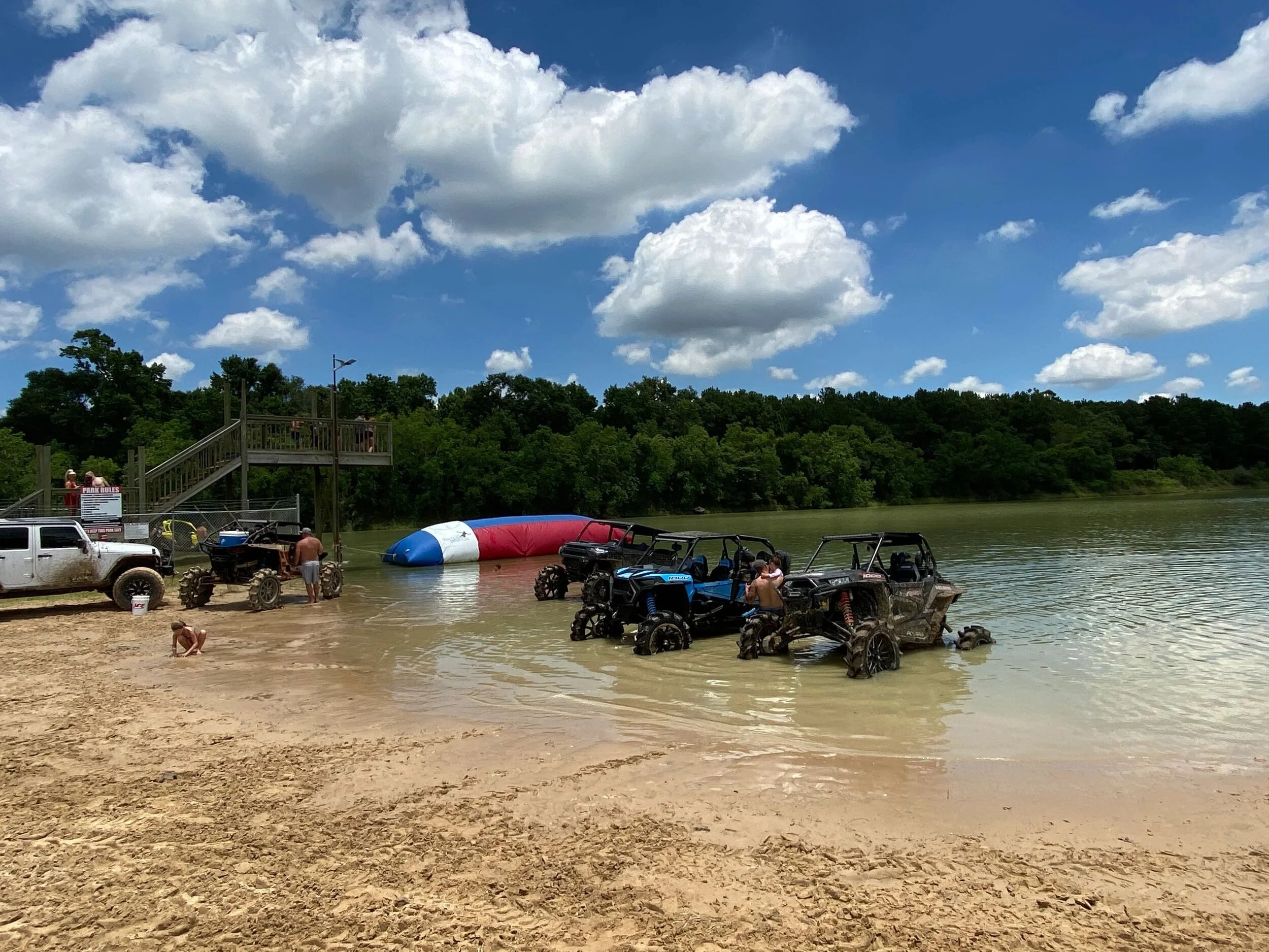 People enjoying a day at a lakeside beach with off-road vehicles, a colorful inflatable water toy, and a boat lift. Some people are on the sandy beach and others near the water, with a green tree-lined shoreline and a partly cloudy sky overhead.