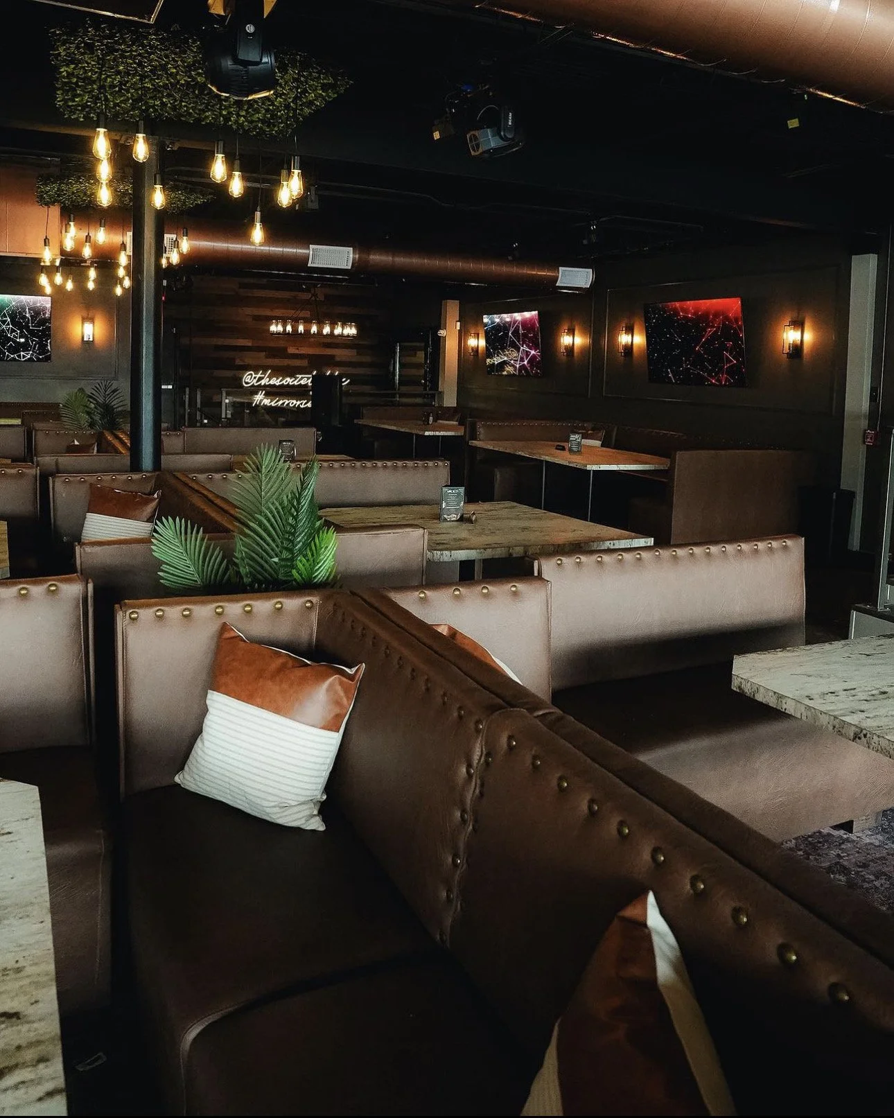 Interior of a modern restaurant or lounge with brown leather booths, throw pillows, wall-mounted televisions displaying digital artwork, hanging Edison bulb lights, and potted plants.