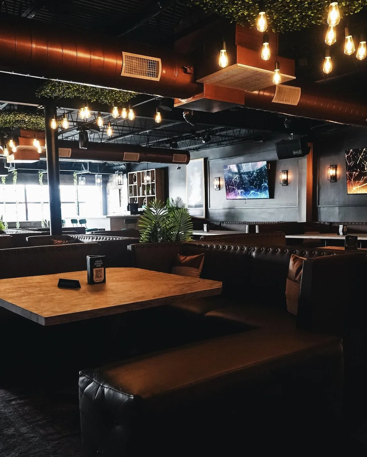 Empty modern restaurant or bar with dark furniture, hanging Edison-style lightbulbs, large windows, and electronic screens on the wall.