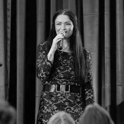 Woman in lace dress speaking into a microphone on stage at Renaissance Newport Beach Hotel, with audience seated in front of her.