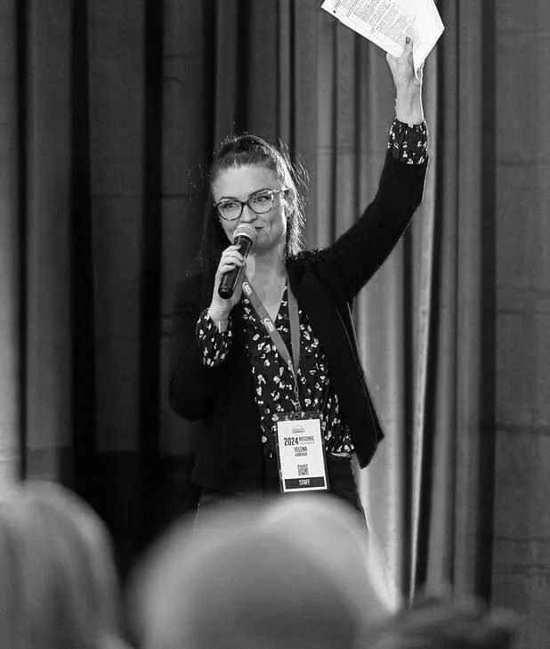A woman with glasses and dark hair, wearing a black blazer and black patterned blouse, holds a microphone in one hand and a paper in the other, standing on stage in front of blue curtains, speaking to an audience.