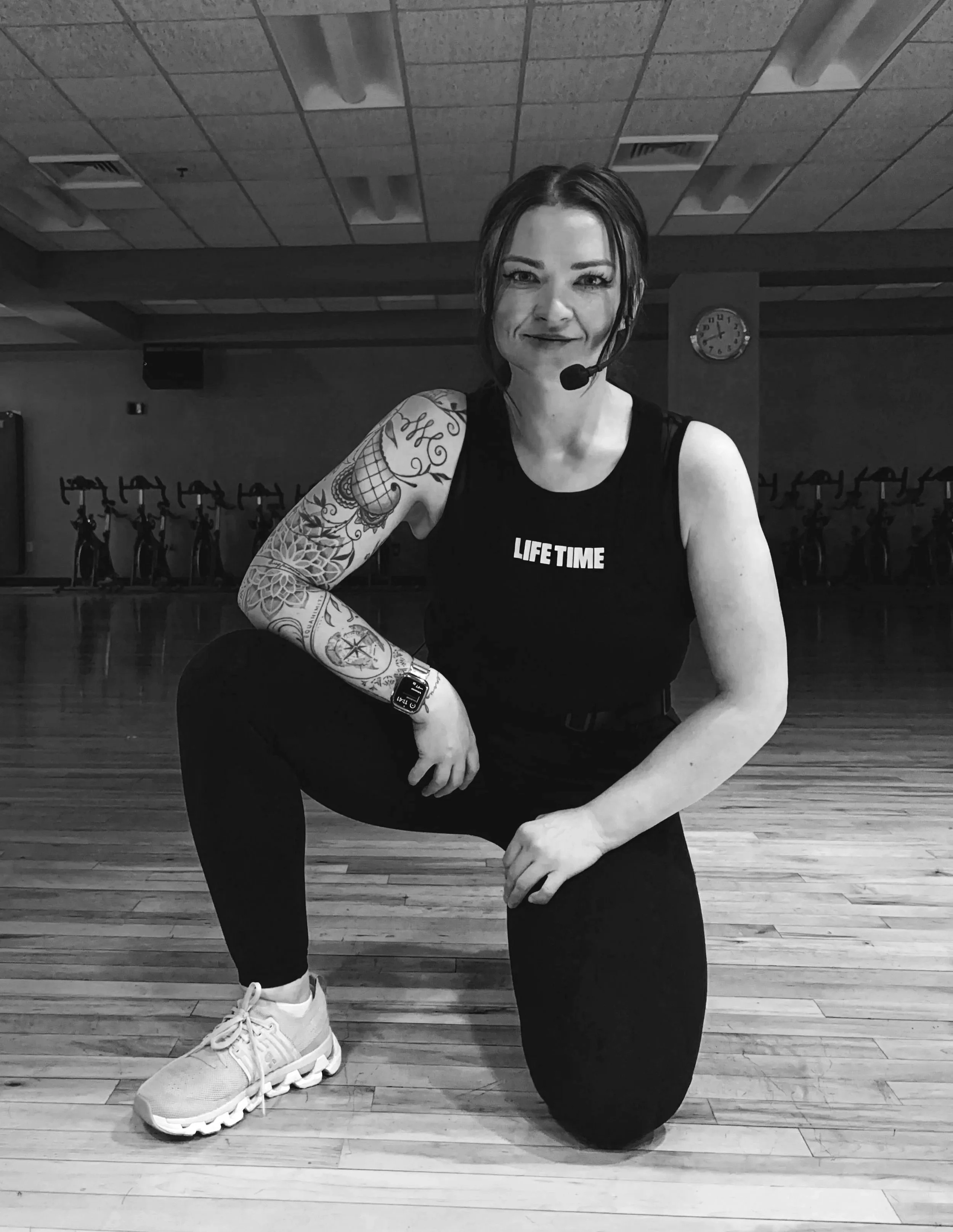 A woman with tattoos on her arm and wearing a black tank top with the words "LIFE TIME" is in a gym, kneeling on one knee and smiling at the camera. She has dark hair, a headset microphone, and is wearing white athletic shoes. The gym has a wooden floor, exercise bikes in the background, and a clock on the wall.