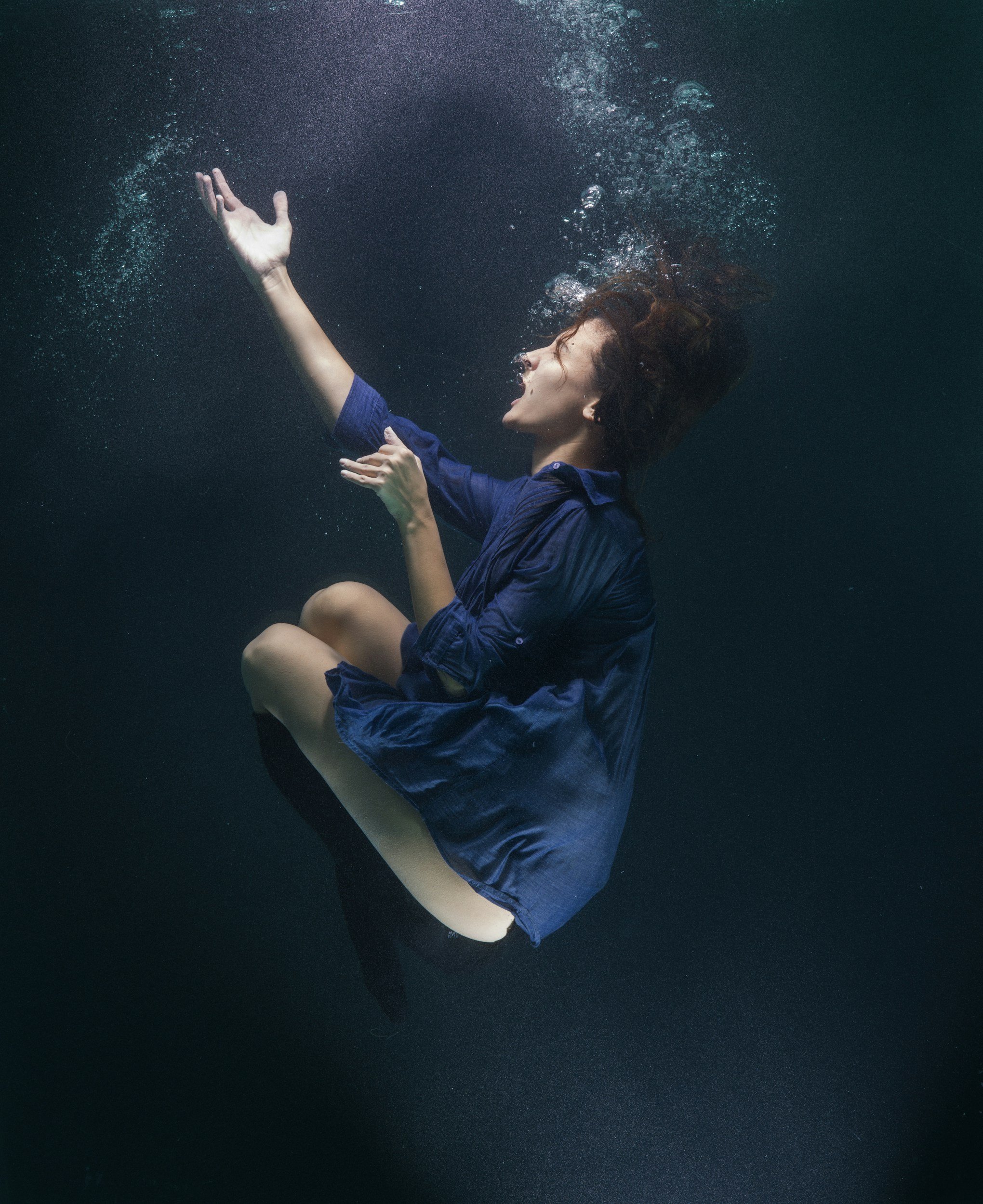 A woman in a blue shirt submerged underwater, with her head tilted back, eyes closed, and one arm raised, creating bubbles around her.