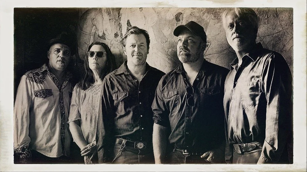 Group of five people standing side by side, in black and white photo, with a wall and artwork in the background.