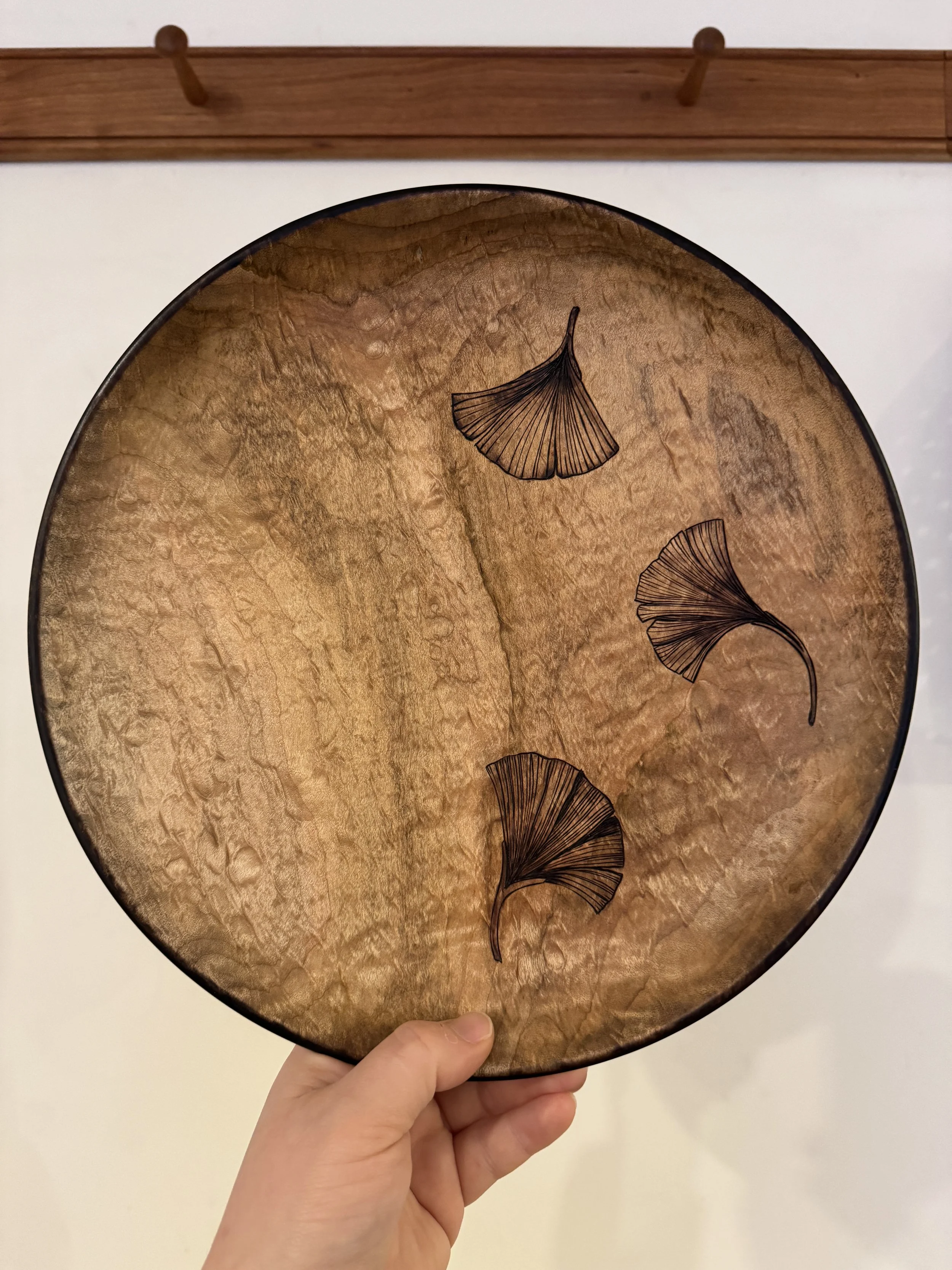 Hand holding a decorative circular tray with black ginkgo leaf illustrations on a textured gold background and a wooden wall with hooks in the background.
