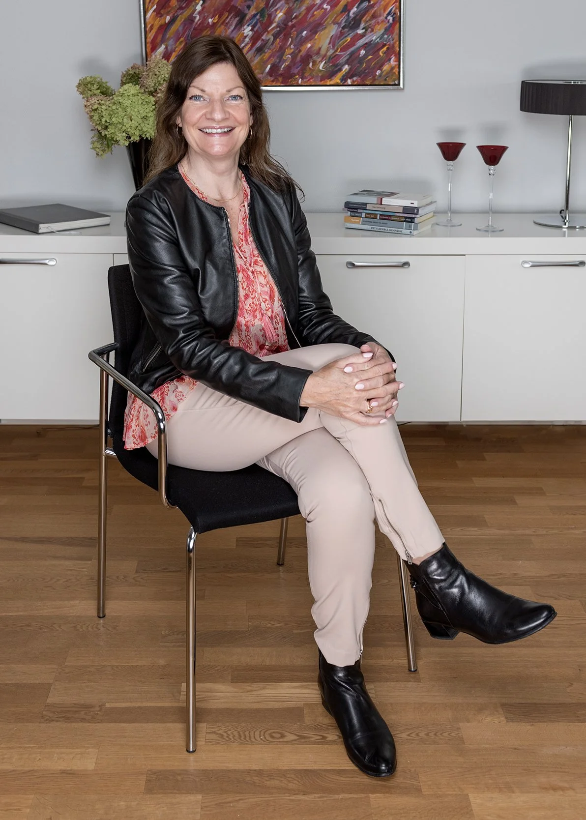 A woman sitting on a black chair in an office, smiling, crossing her legs, wearing a black leather jacket, pink lace blouse, beige pants, and black boots. Behind her is a white cabinet with a small stack of books, two red glasses, a black desk lamp, and a plant with green flowers. A colorful abstract painting hangs on the wall.