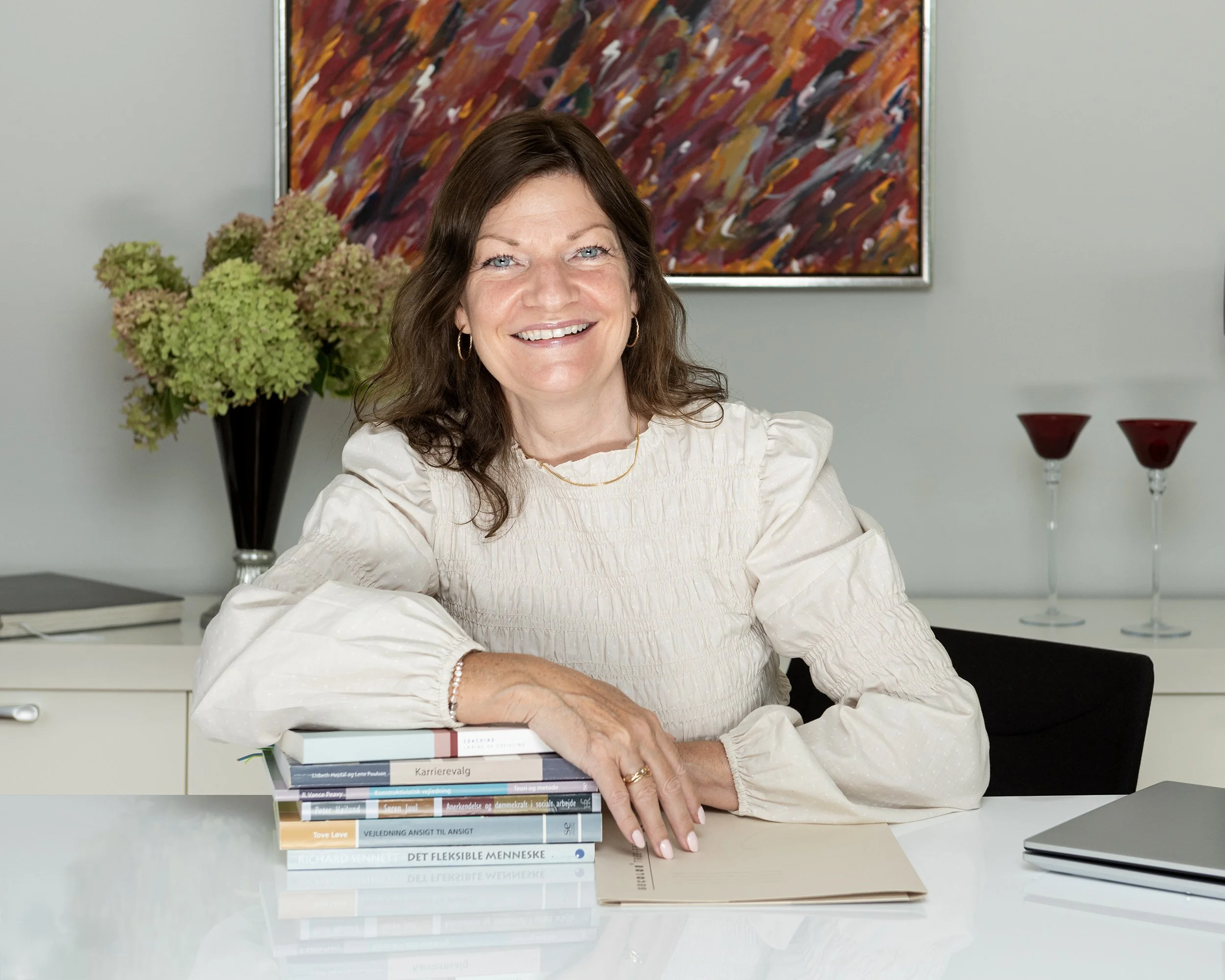 A smiling woman with shoulder-length brown hair and blue eyes, sitting at a white desk with a stack of books, a closed laptop, and a brown folder. Behind her is a large colorful abstract painting, a black vase with green hydrangeas, and three red candle holders with clear stems. The background is a plain, light-colored wall.