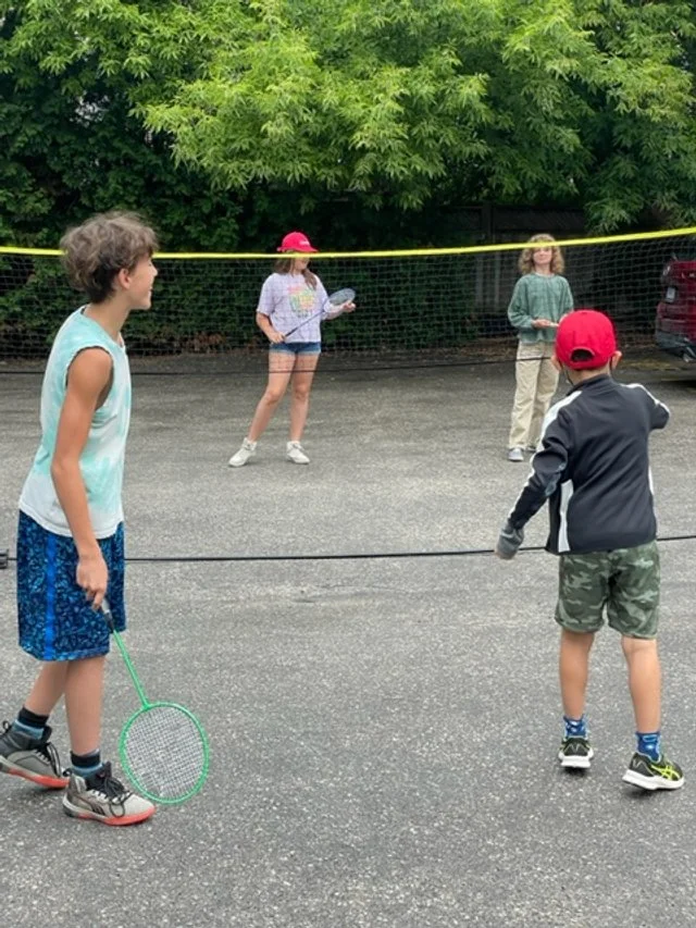 Children playing badminton outdoors on a paved area with a green, leafy background.