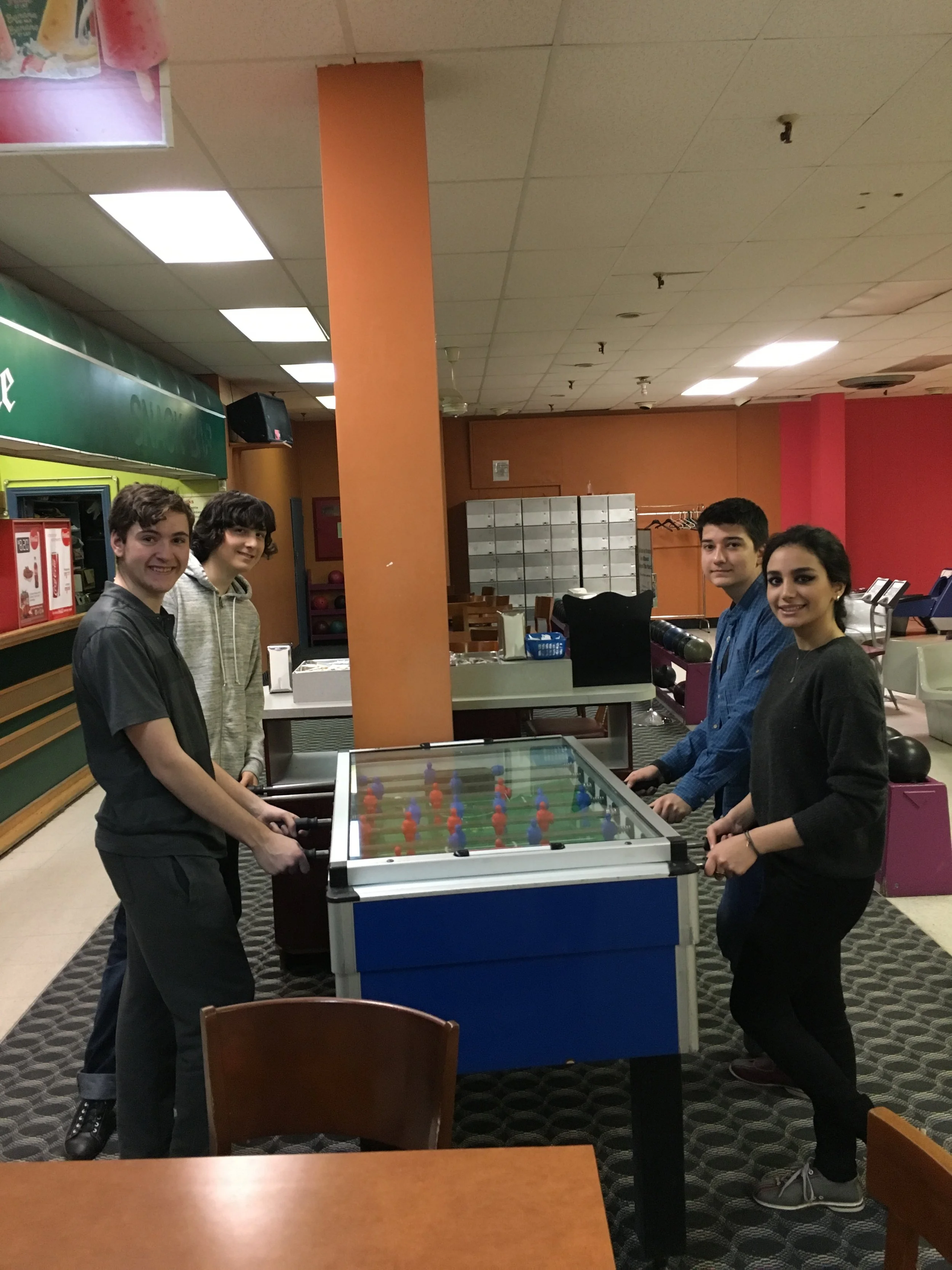 Five young people standing around an air hockey table in a bowling alley or recreation center, smiling at the camera.