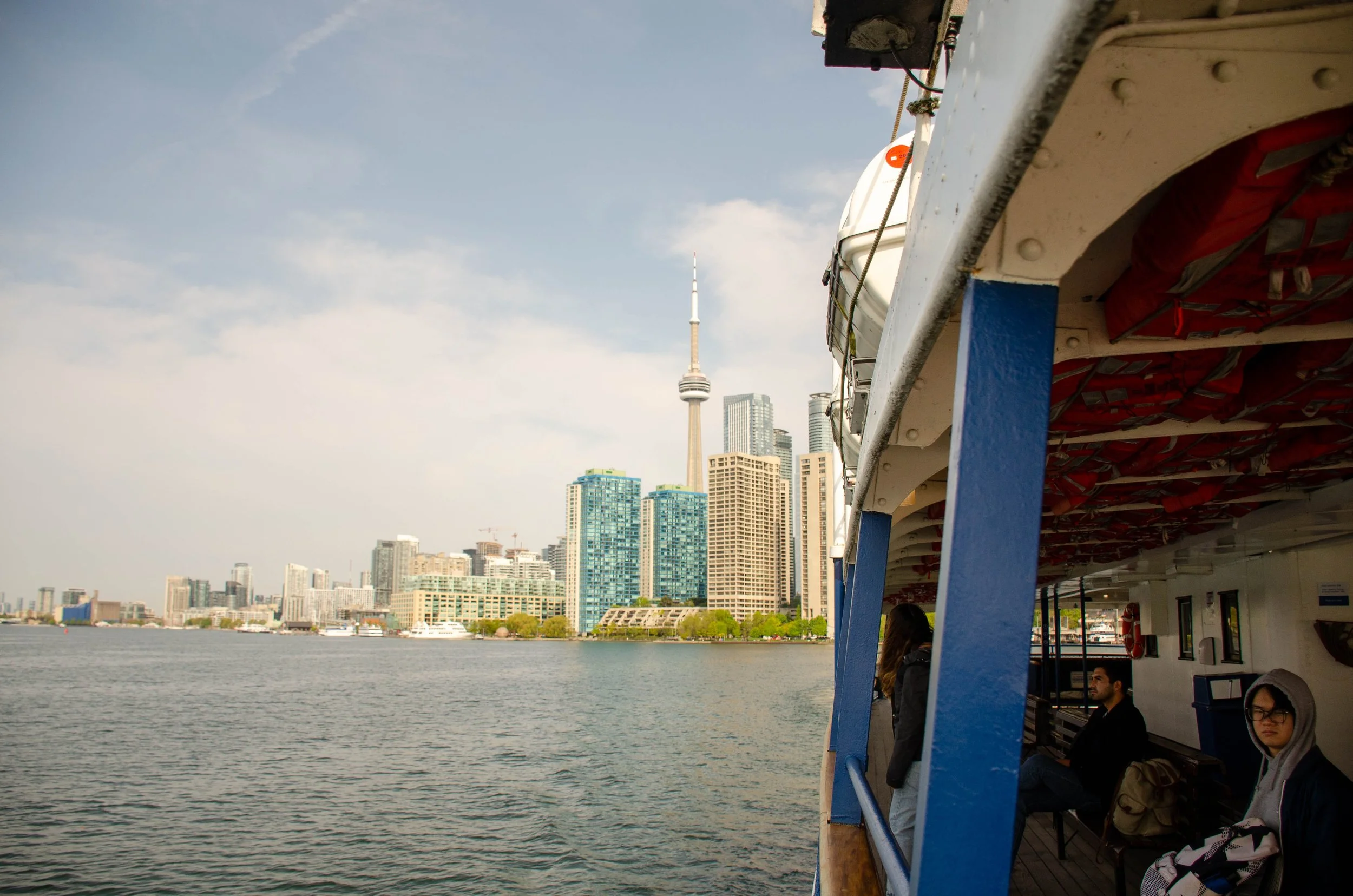 View of Toronto skyline from a ferry, with the CN Tower prominent in the background, and passengers seated on the ferry's deck.