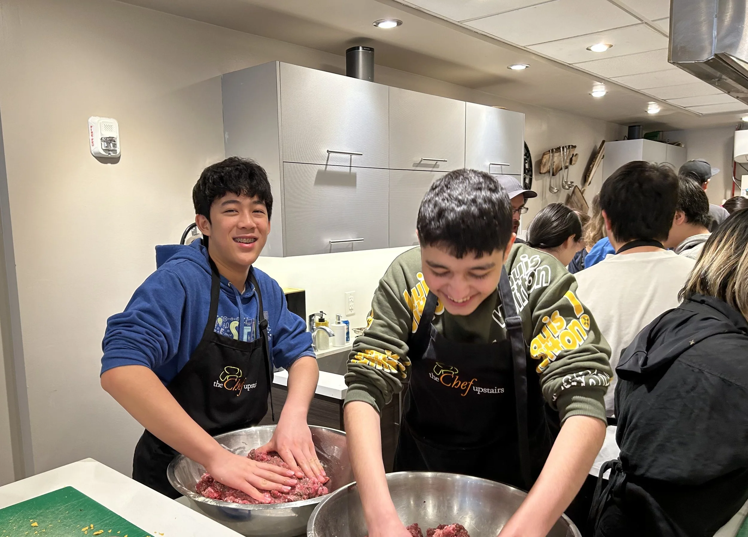 Young boys smiling and mixing ground meat in large bowls in a kitchen, wearing aprons, with a group of people in the background.