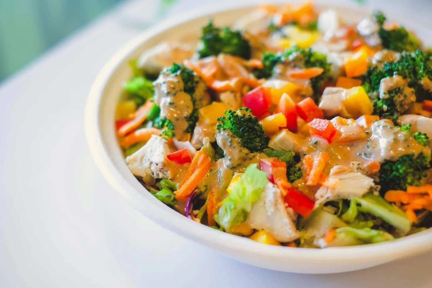 Close-up of a colorful chicken and vegetable salad with broccoli, red and yellow peppers, carrots, and lettuce in a white bowl.
