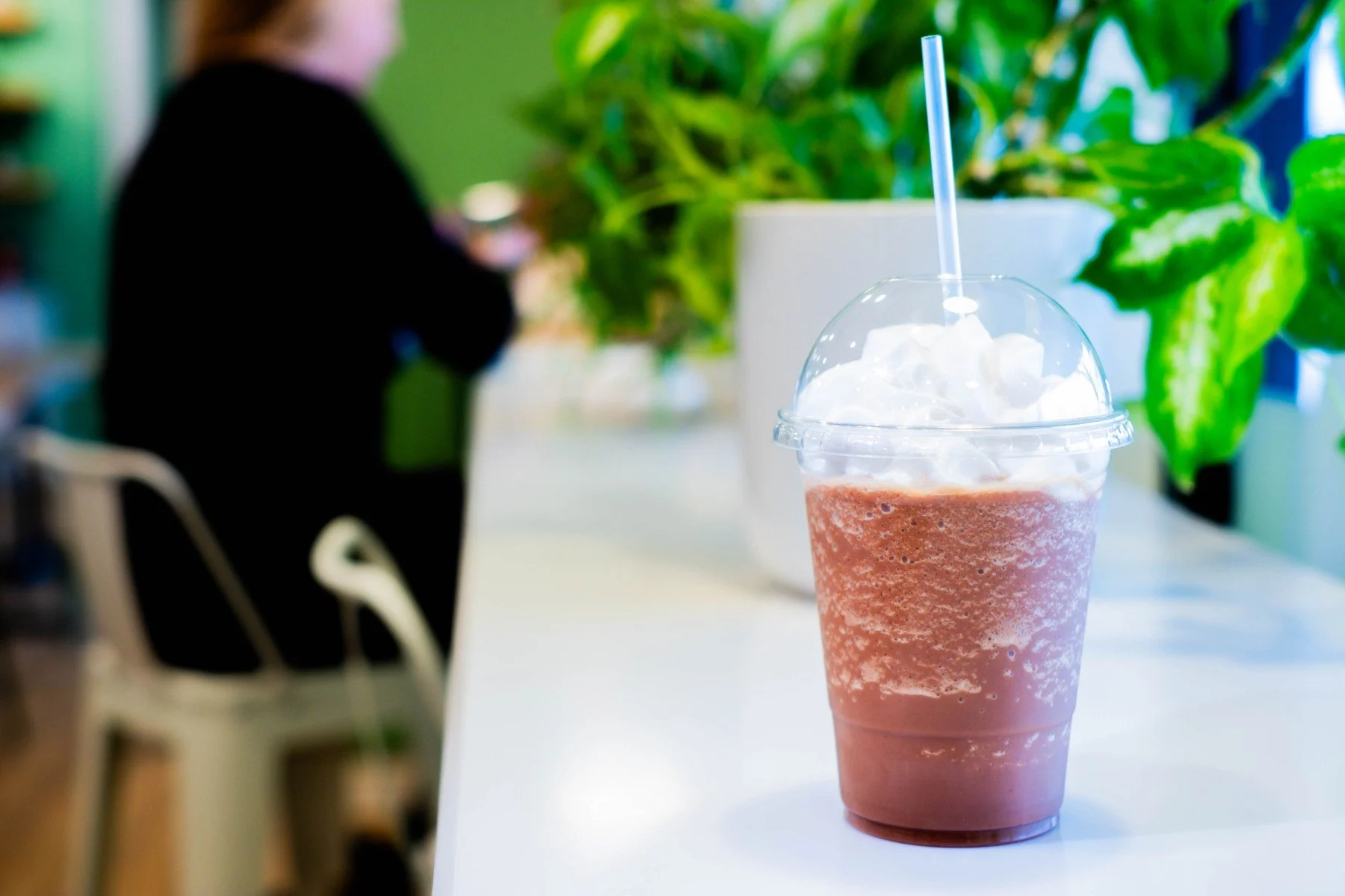 a frappe or frappuccino in a plastic cup and lid with a straw, with a person in the background sitting in front of a window