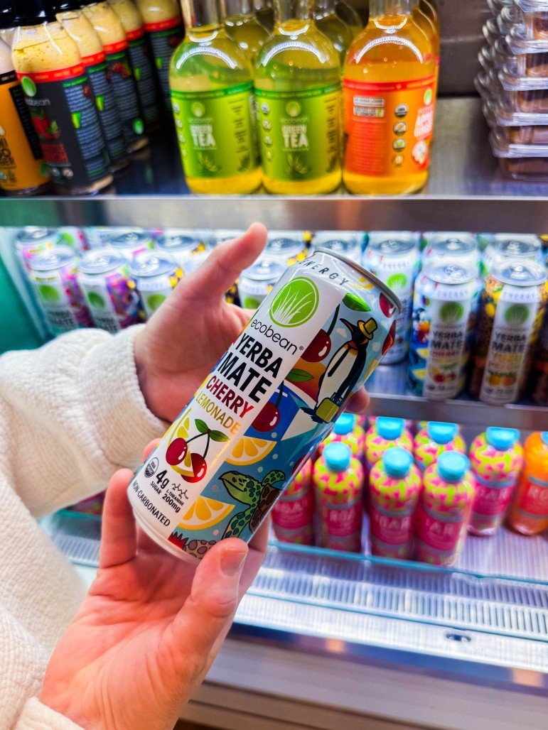 A person holding a can of Ecobean Yerba Mate Cherry Lemonade in front of a refrigerated store shelf stocked with energy drinks and flavored waters.
