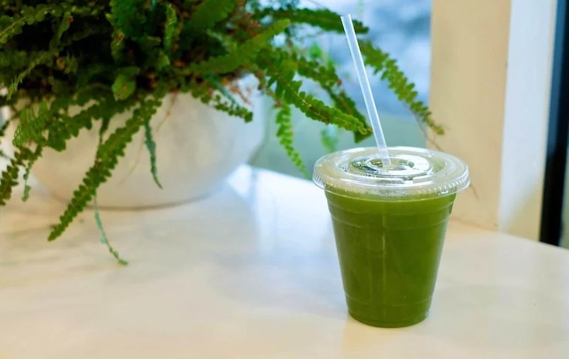 A clear plastic cup with a green juice and a straw on a white surface, with a potted fern plant in the background near a window.