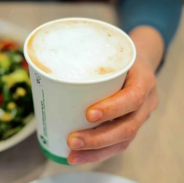 Person holding a white disposable cup with a frothy coffee beverage, possibly a latte or cappuccino.