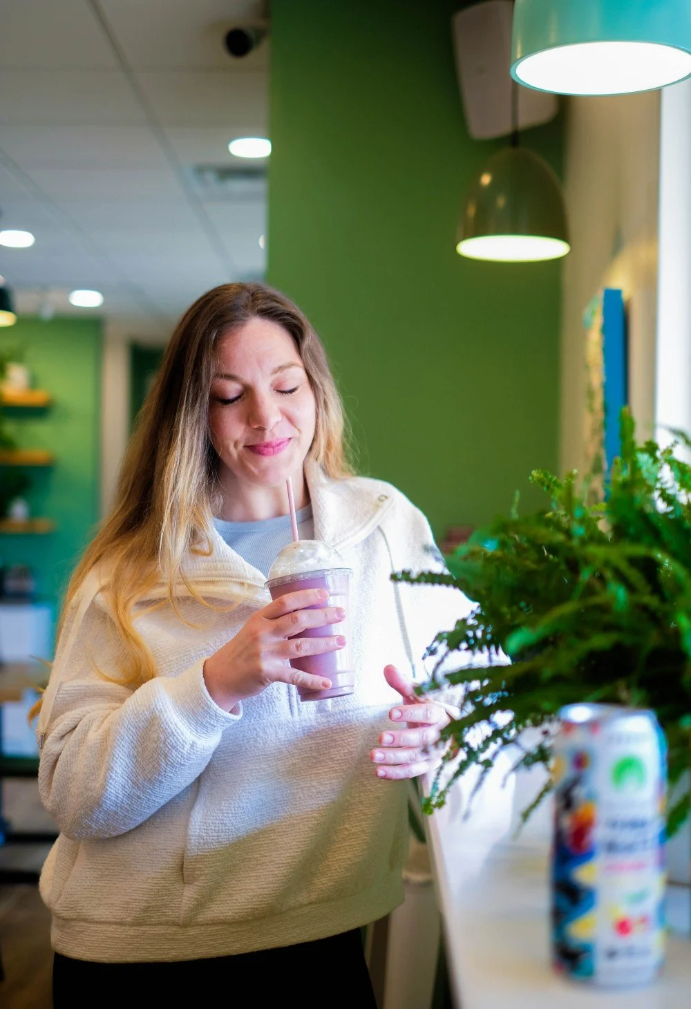 A woman with long blonde hair wearing a beige hoodie, holding a purple smoothie with a straw, smiling with eyes closed in a modern green-themed cafe.
