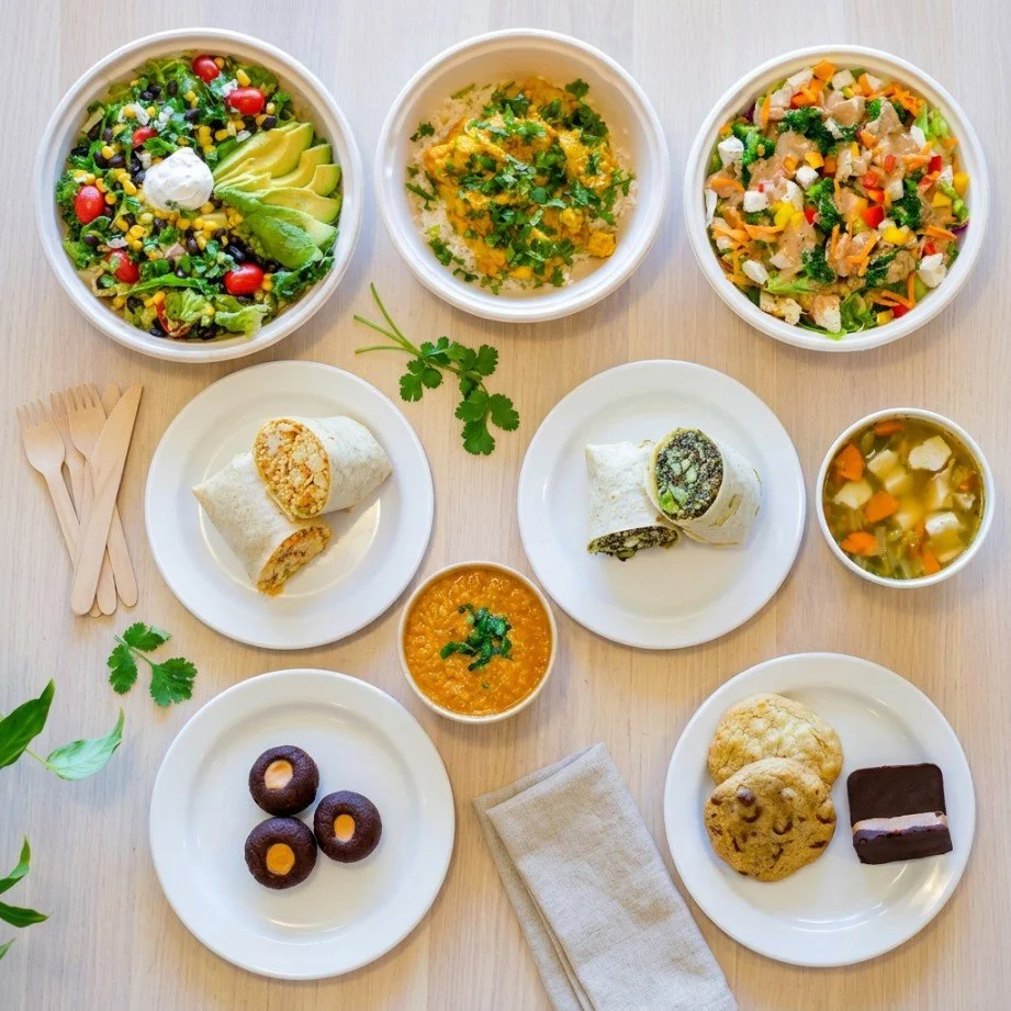 An assortment of dishes including salads, rice, beans, wraps, soup, cookies, and chocolates on a light wooden table.