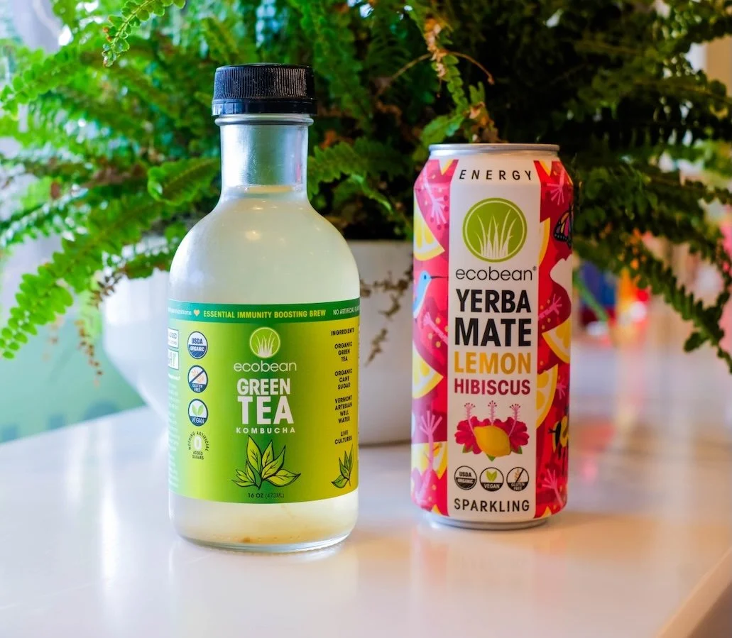 A bottle of Ecobaean Green Tea Kombucha and a can of Ecobaean Yerba Mate Lemon Hibiscus sparkling drink placed on a white surface with a large green fern plant in the background.